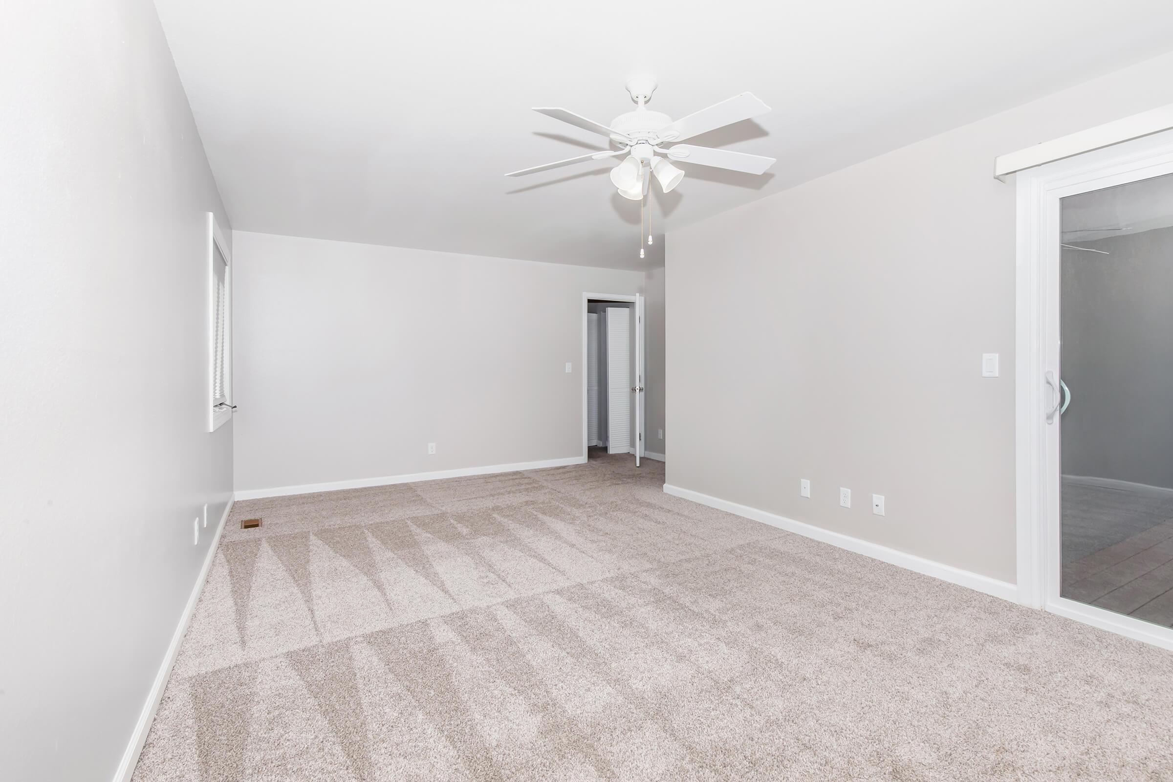 Spacious room with light gray walls and plush beige carpet. A ceiling fan is installed, and there is a large sliding glass door leading to a patio area. The room features two doorways leading to other spaces, creating a bright and open atmosphere.