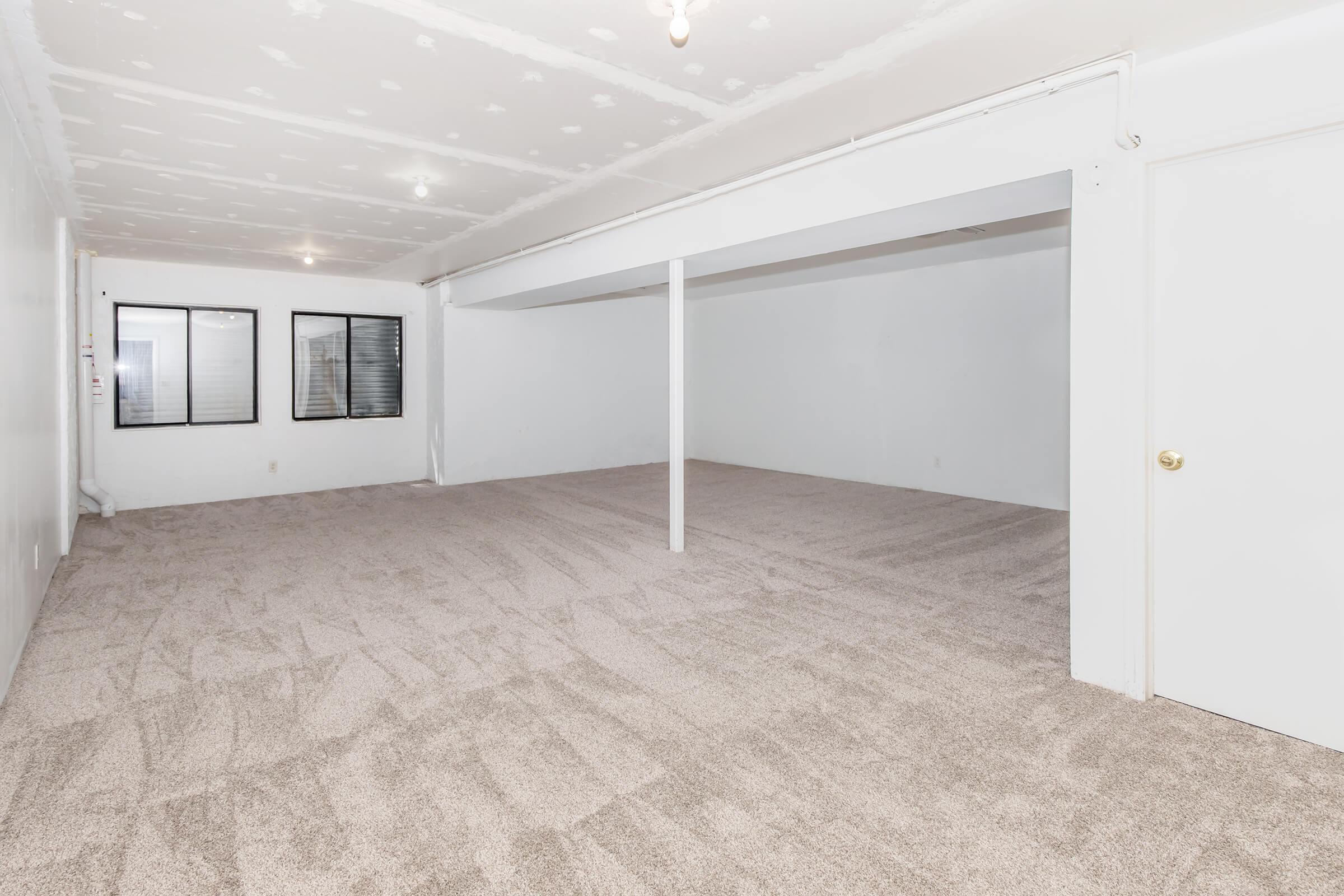 A spacious, empty room with freshly carpeted flooring. The walls are painted white, and there are two windows with black frames on one side. A central support column is present, and the room has a well-lit ceiling with exposed bulbs. Overall, the space appears clean and ready for use.