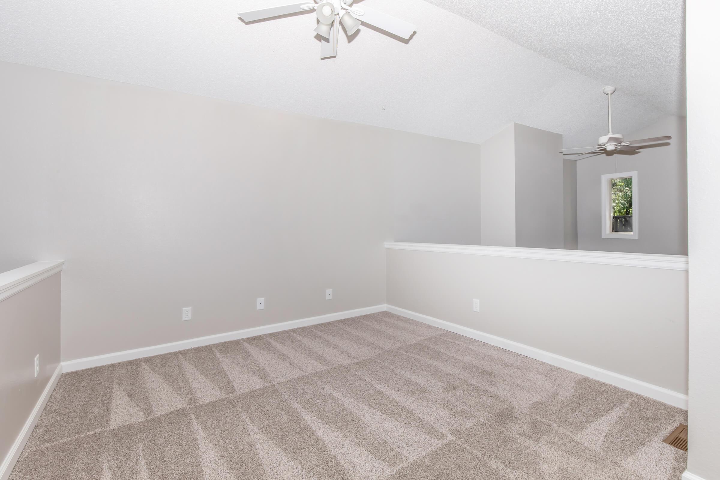 A spacious, softly lit interior room featuring light gray walls and plush carpet. The ceiling is adorned with a ceiling fan, and there is a small ledge that overlooks a lower area. The room has a clean and minimalistic design, creating a cozy and inviting atmosphere.