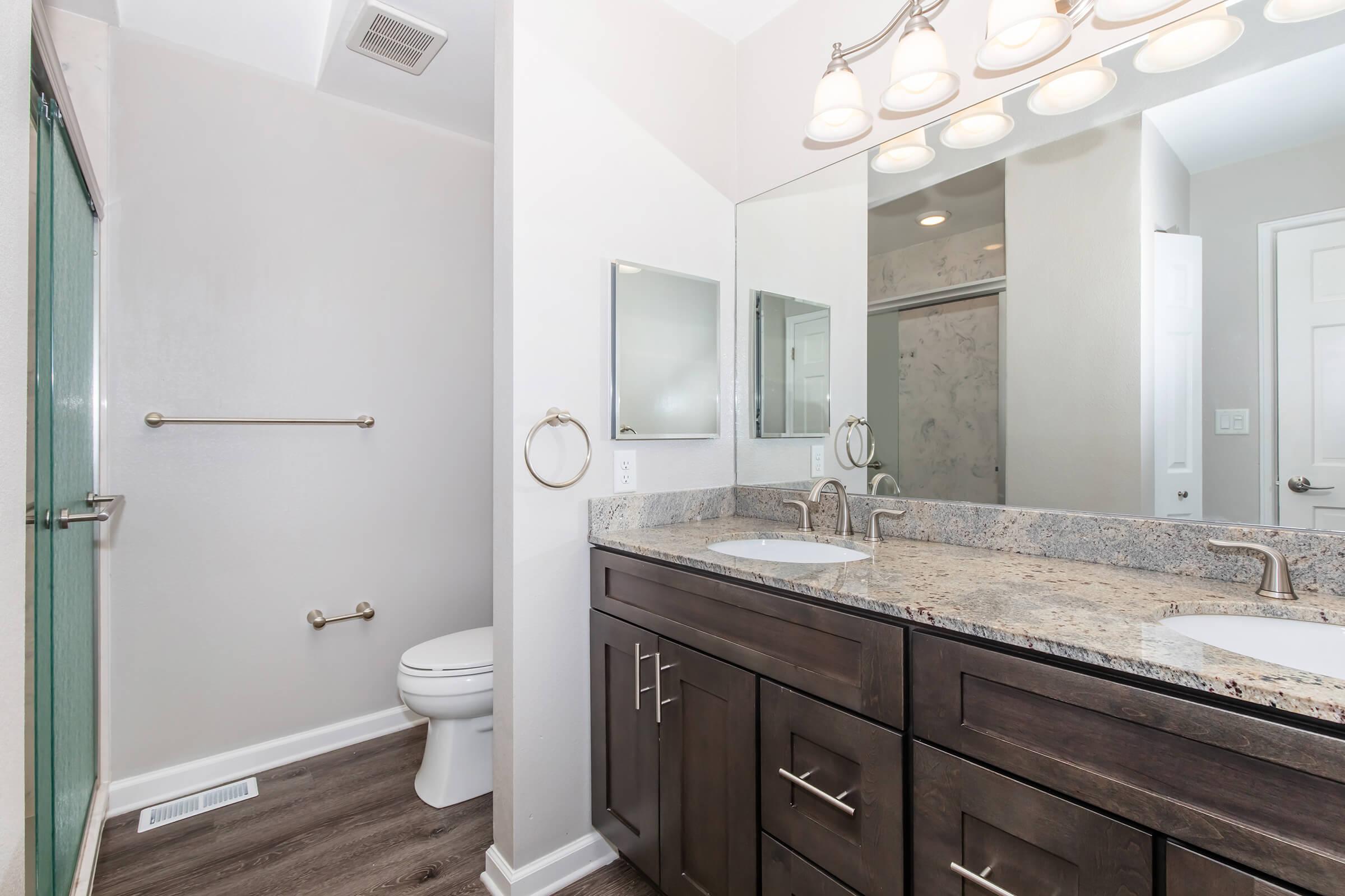 A modern bathroom featuring a double sink vanity with granite countertops, a large mirror, a shower area with a glass door, a towel bar, and a toilet. The walls are painted light gray, and the flooring is a dark wood laminate. Bright lighting fixtures hang above the vanity.