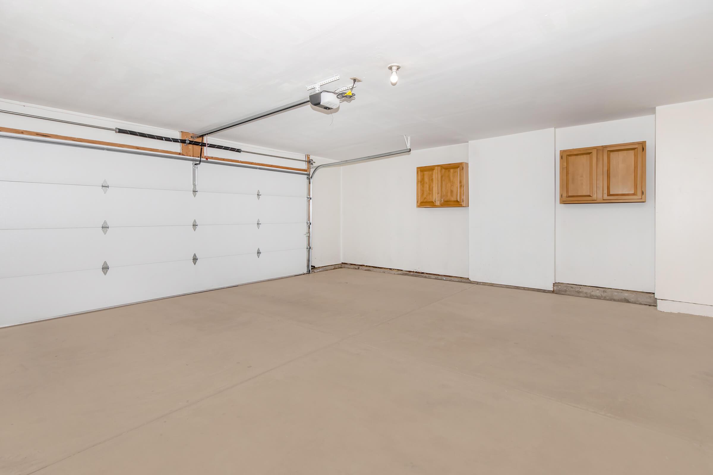 A spacious, empty garage with a white garage door, concrete floor, and light-colored walls. There are wooden cabinets mounted on the wall, providing storage space. The room has good lighting from an overhead fixture, creating a bright and clean environment.