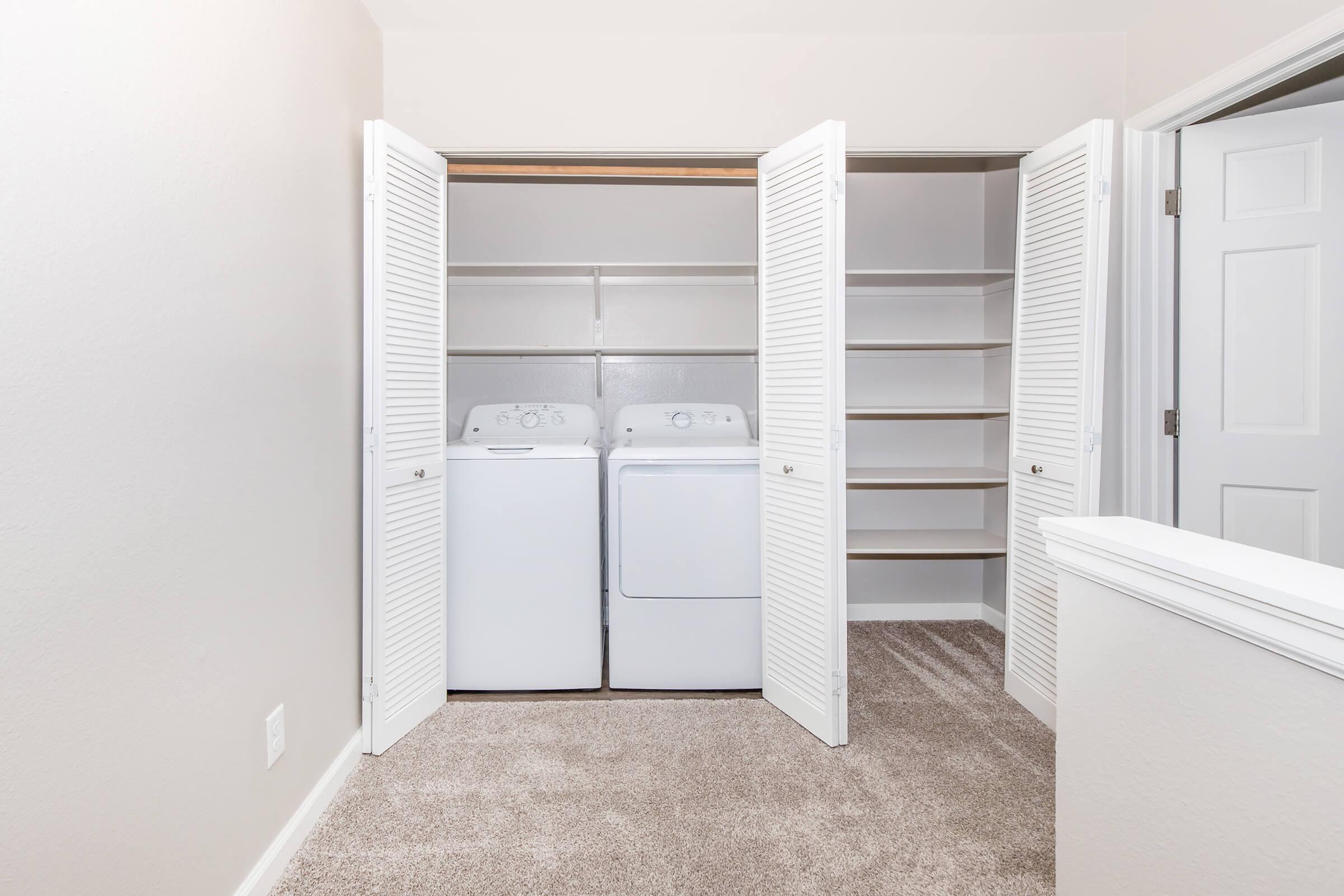 A clean laundry room with a stacked washer and dryer enclosed by white bi-fold doors. The adjacent wall features open shelves for storage. The space has light-colored walls and carpet, creating a bright and organized environment.