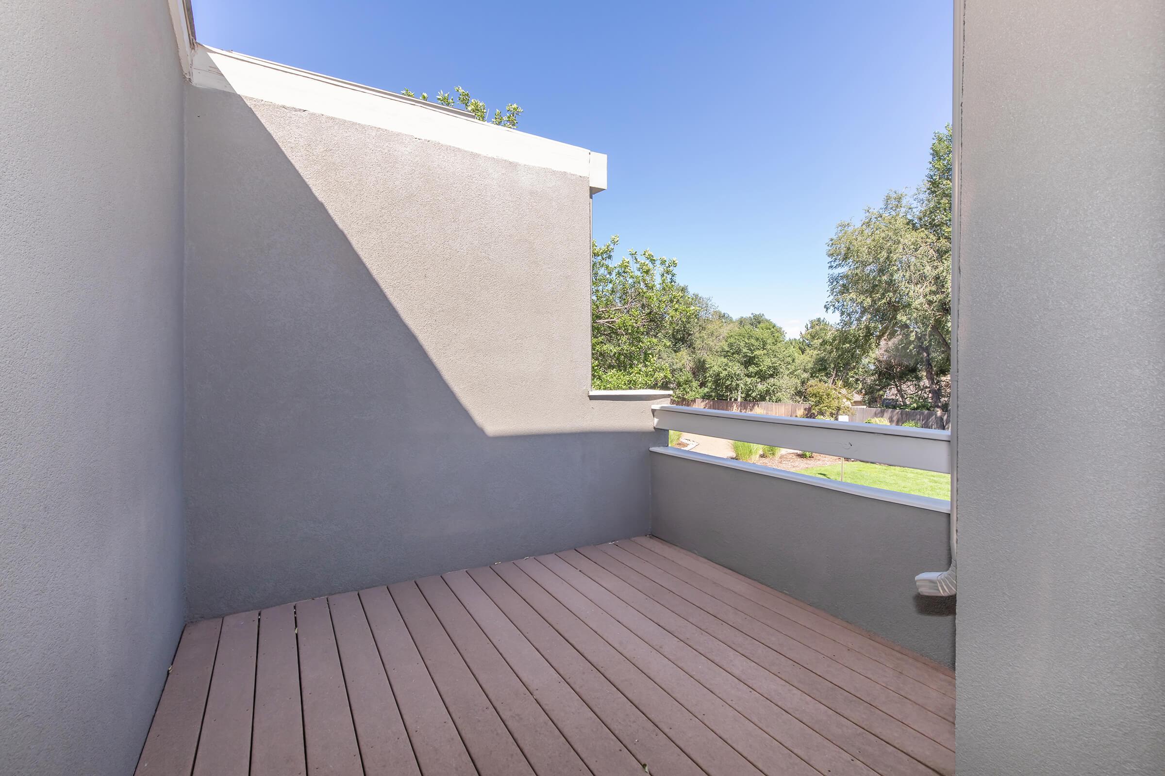 A small balcony or patio area with wooden flooring, surrounded by light gray walls. The sky is clear and blue, with greenery visible in the background, creating a serene outdoor space for relaxation.