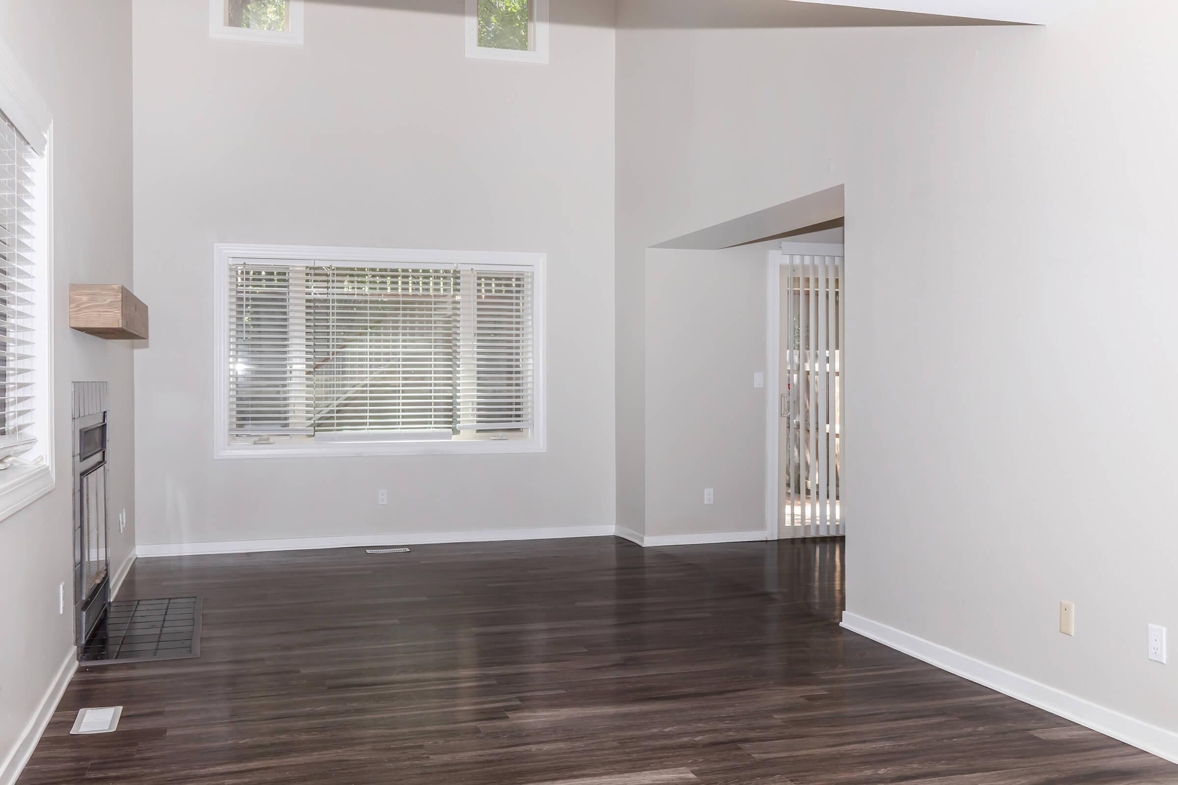 A spacious, empty living room with light-colored walls and dark hardwood flooring. Large windows with blinds allow natural light in, and a fireplace is positioned on one wall. An archway leads to another area, and a door with a security screen is visible in the background.