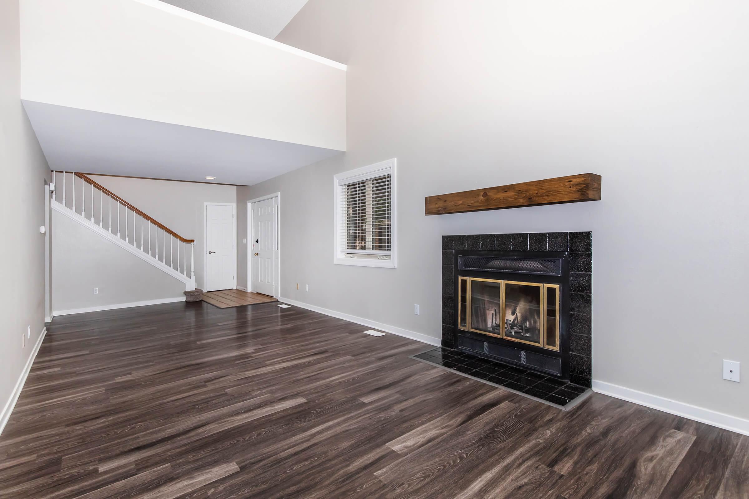Spacious interior of a living room featuring hardwood floors, a black fireplace with a wooden mantle, and large windows. A staircase is visible in the background, leading to an upper level. The walls are painted in a light color, creating an airy and inviting atmosphere.