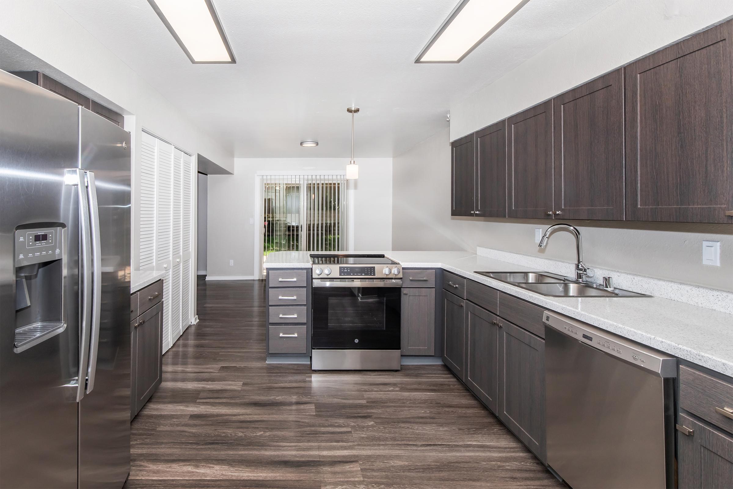 Modern kitchen featuring dark wooden cabinets, stainless steel appliances, and a large island. The space includes a refrigerator, oven, dishwasher, and a double sink. There's a window with vertical blinds allowing natural light, and the flooring is dark wood, adding warmth to the contemporary design.