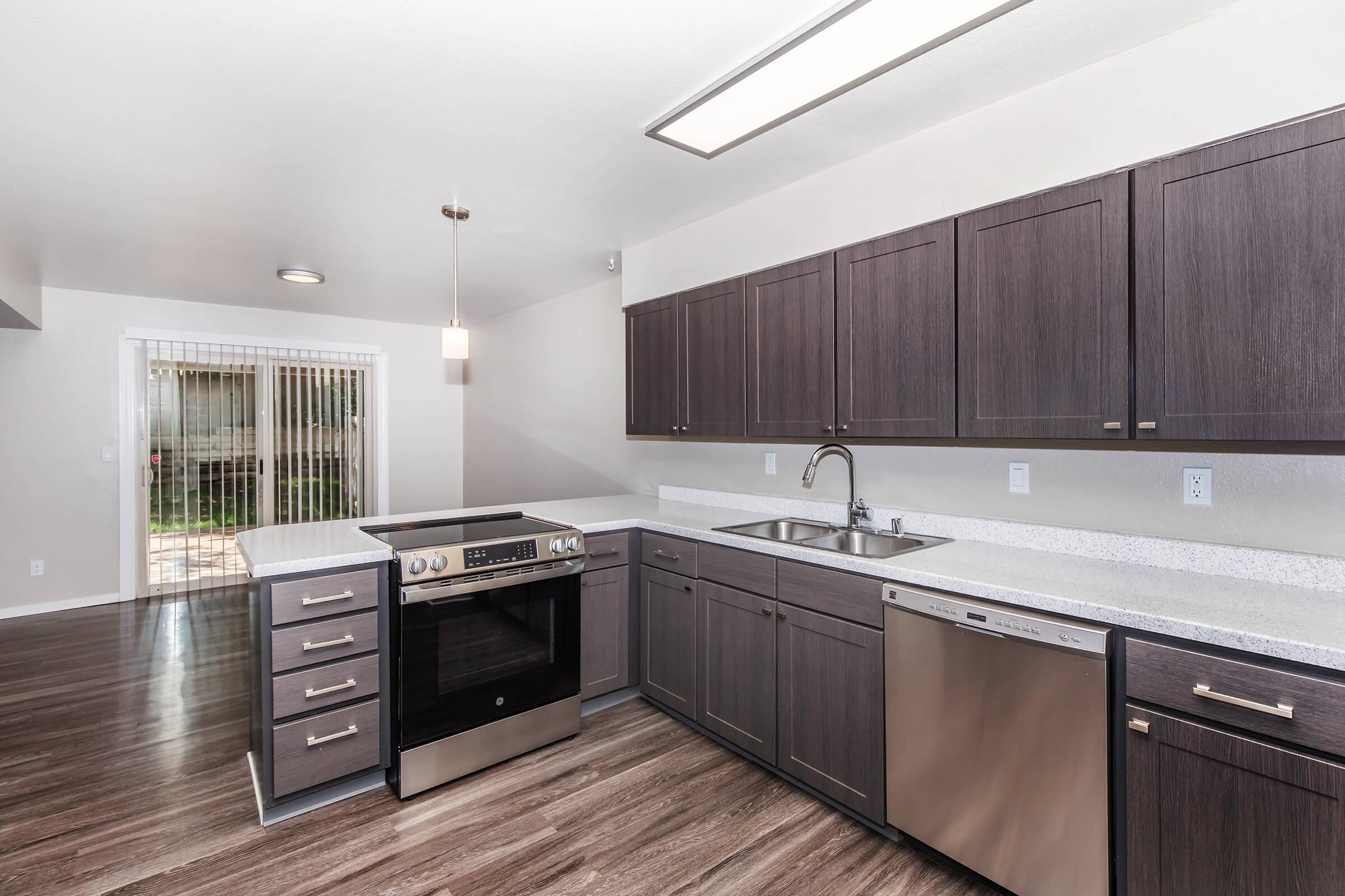 Modern kitchen featuring dark wooden cabinetry, stainless steel appliances including an oven and dishwasher, and a white countertop. The space has a contemporary light fixture and is open to a bright dining area with sliding glass doors leading to an outdoor patio. The floor has a sleek, dark finish.