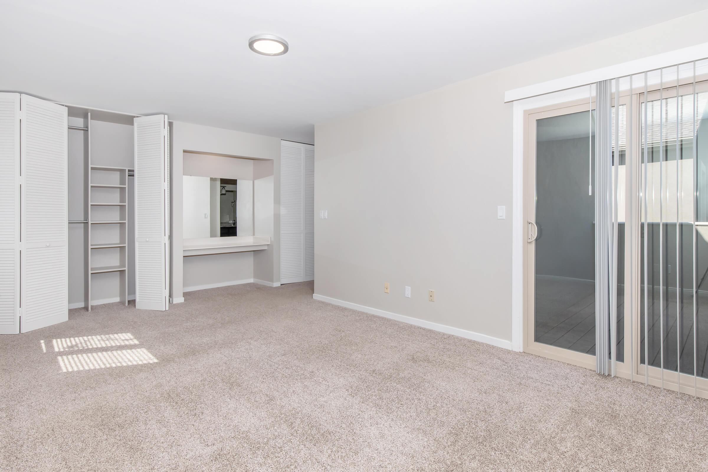 A spacious, empty room with light-colored walls and carpeted flooring. There are shutter doors on the left leading to a closet space and a vanity area. On the right, there's a sliding glass door that leads outside, allowing natural light to flood the room.