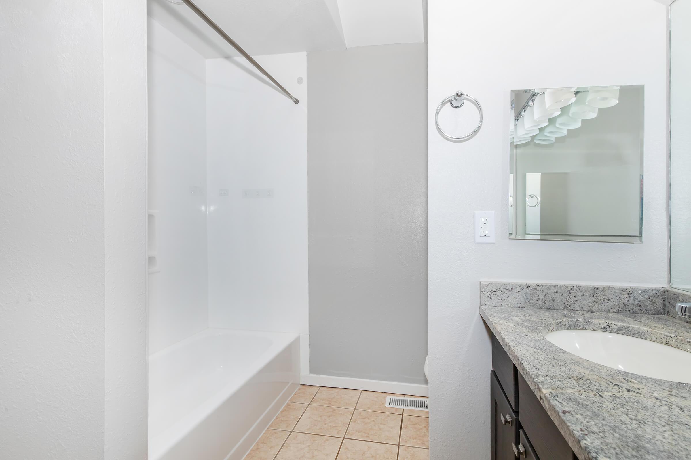 A clean bathroom featuring a white bathtub and shower, light gray walls, and a dark wood vanity with a granite countertop. A mirror with multiple light fixtures is mounted above the sink, and beige floor tiles complete the design. The space is bright and well-lit.