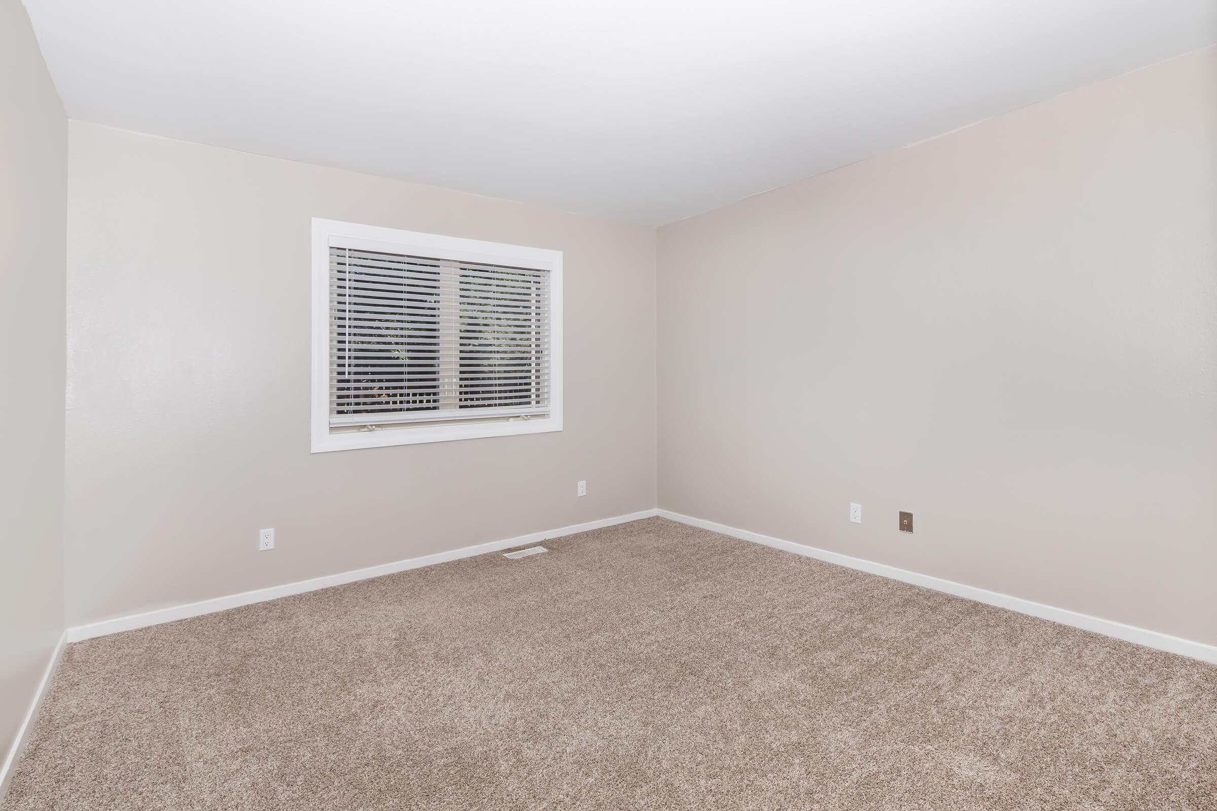 Empty room with light beige walls and a window featuring white blinds. The floor is covered in beige carpet. There are no furniture or decorations present, creating an open and spacious feel.