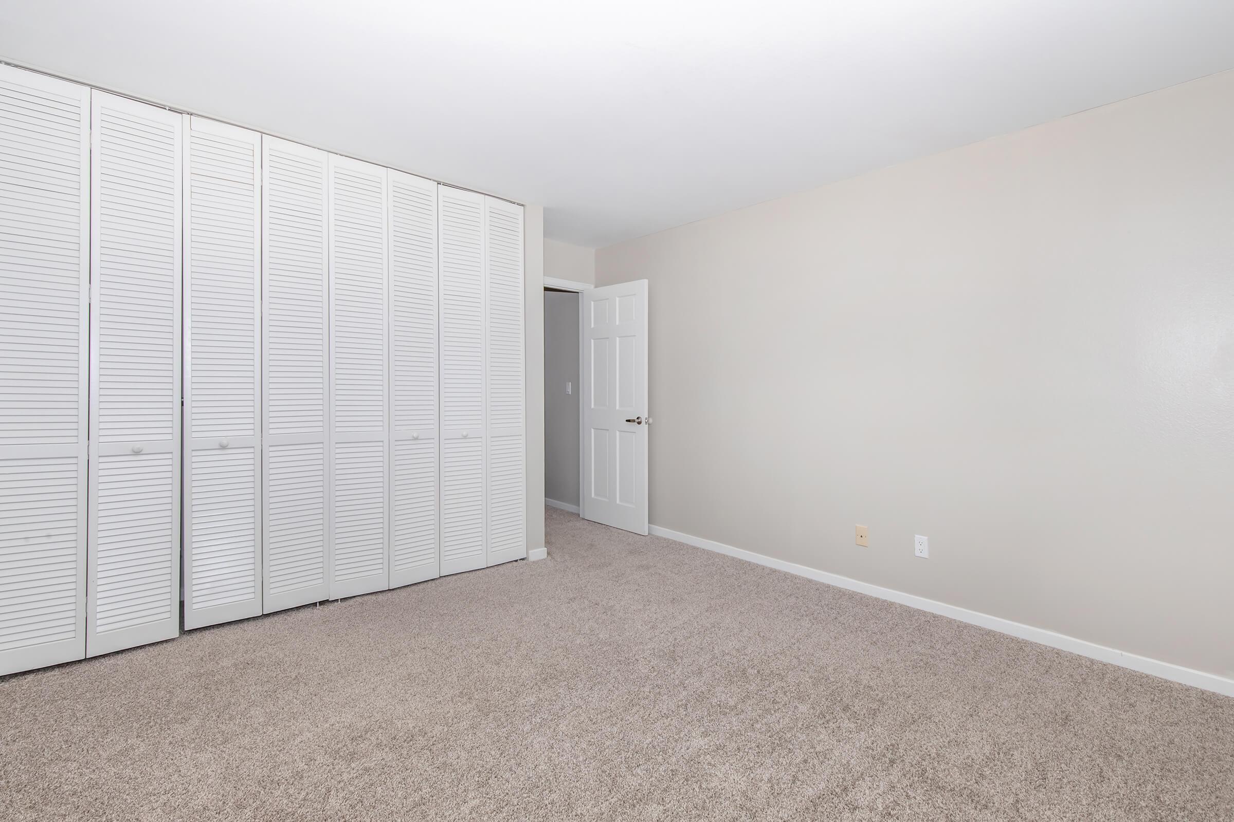 A spacious, empty room with light beige walls and plush carpet. One wall features white, louvered closet doors, while another wall has a closed door leading to another space. Natural light brightens the room, creating a clean and airy atmosphere.