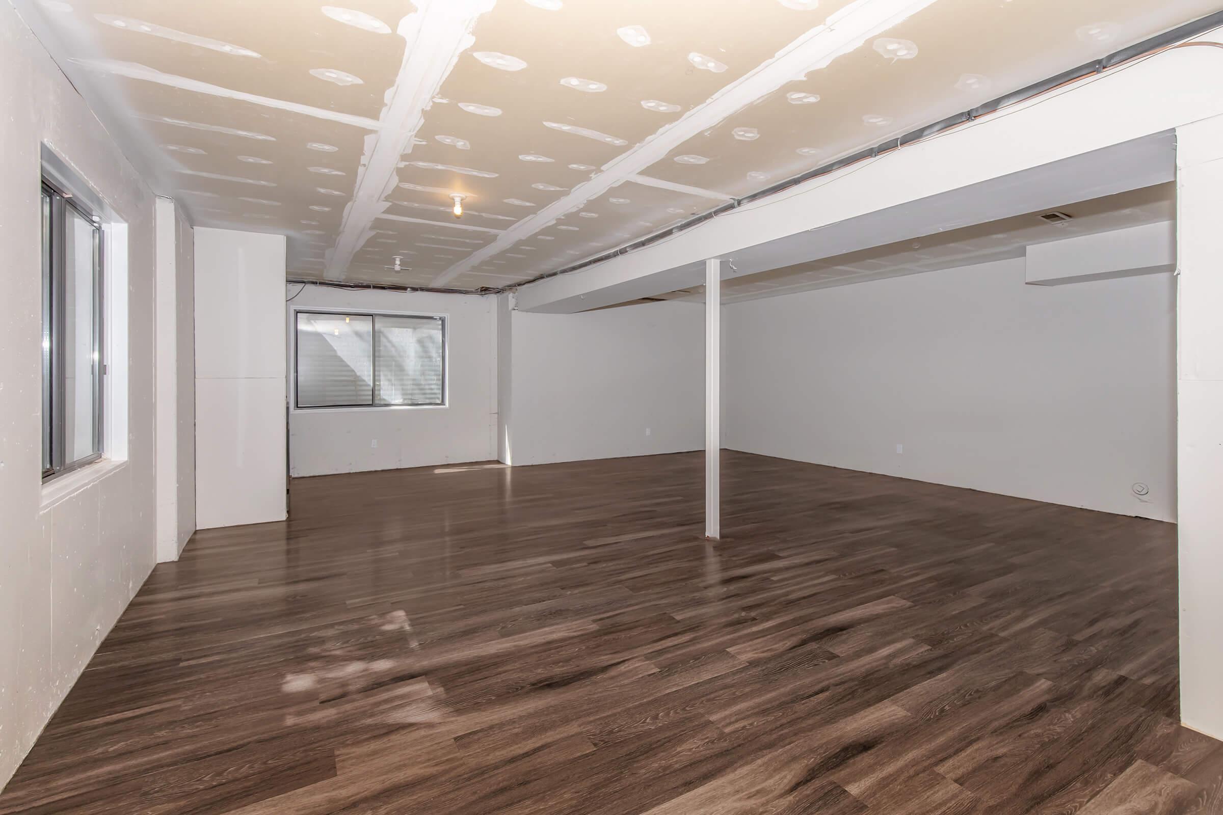 A spacious, empty basement room with light-colored walls and a smooth, polished wooden floor. There is a window on one wall allowing natural light to enter, and the ceiling features exposed beams with a drywall finish. The space is clean and ready for customization.