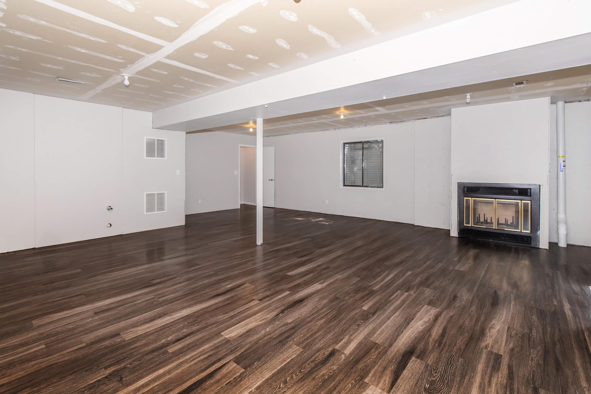 Spacious unfinished basement with a polished wood floor, exposed ceiling, and a gas fireplace against a plain wall. The room features a window and door leading to another area, with drywall ready for finishing touches. Soft lighting from ceiling fixtures adds a warm ambiance to the space.