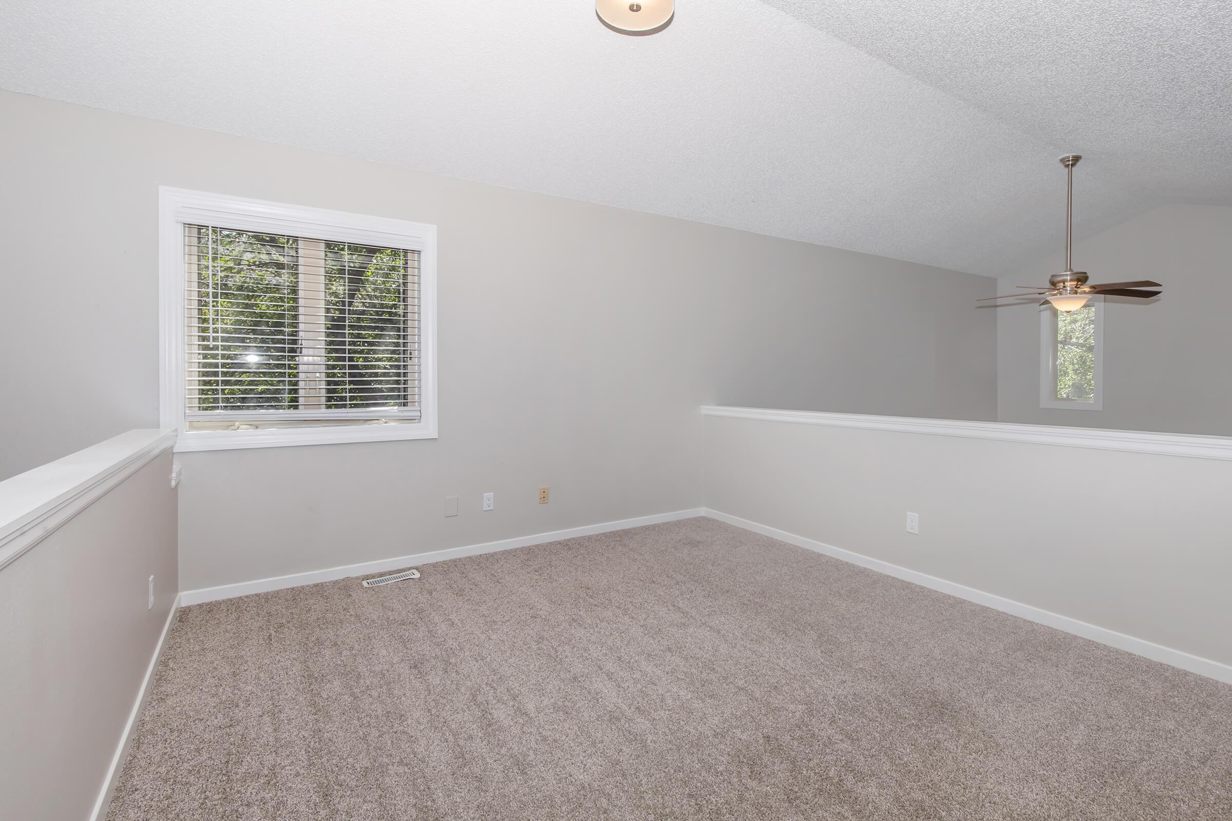 A well-lit, empty room featuring beige walls and carpet, with a ceiling fan and a window allowing natural light. The space has a sloped ceiling, and a railing separates it from an adjacent area, creating an open and airy feel. The overall design is simple and modern.