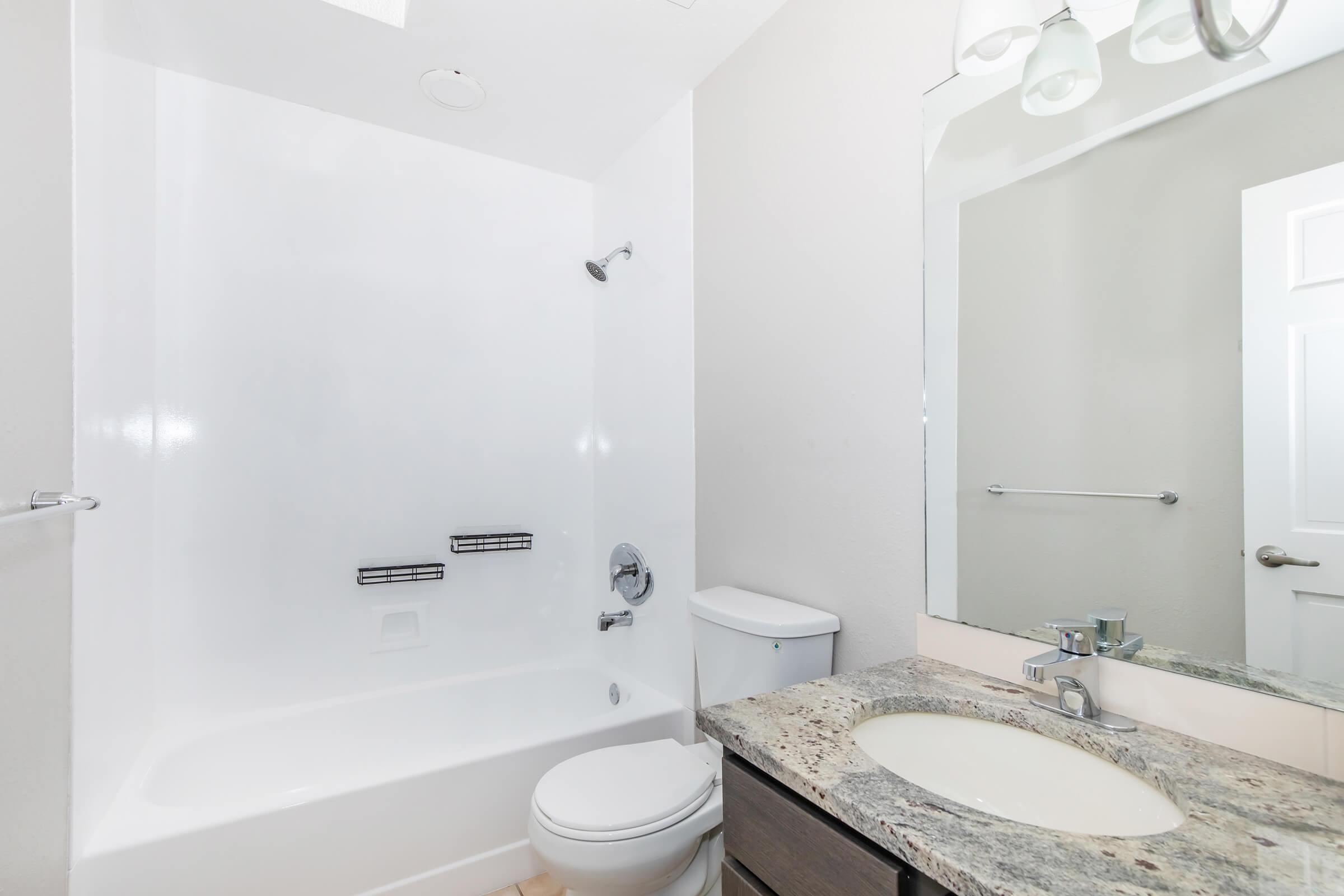 A clean, modern bathroom featuring a white bathtub and shower combination, a toilet, and a granite countertop with a sink. A large mirror hangs above the sink, and there are minimalistic fixtures and a towel bar, creating a bright and spacious appearance. The walls are painted in a light color.