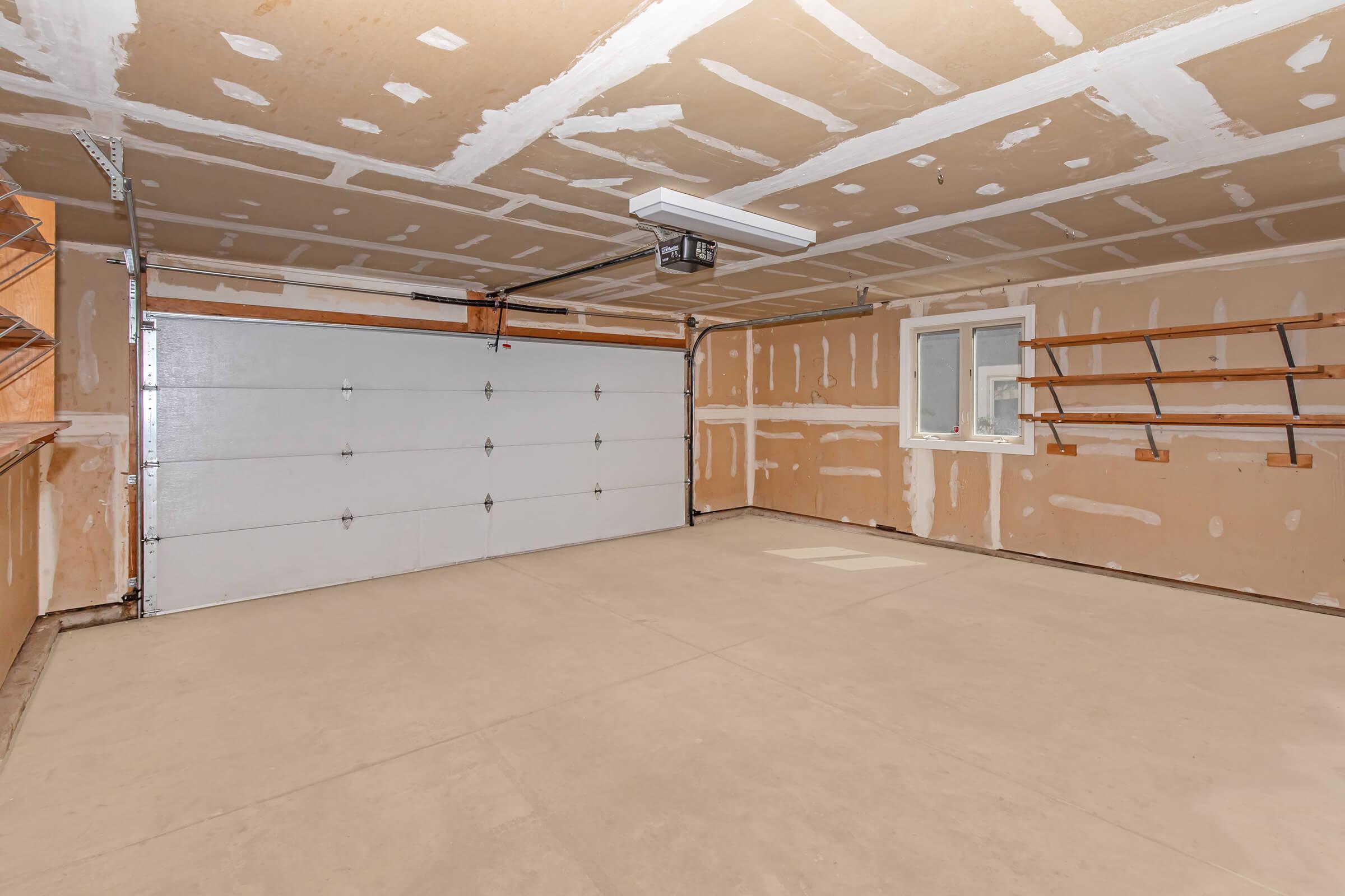 An empty garage featuring a concrete floor, a large white garage door, and unfinished drywall walls with visible tape and joint compound. Shelves are mounted on one side, and a window is present, allowing natural light into the space. The overall atmosphere is clean and organized, ready for storage or workspace use.