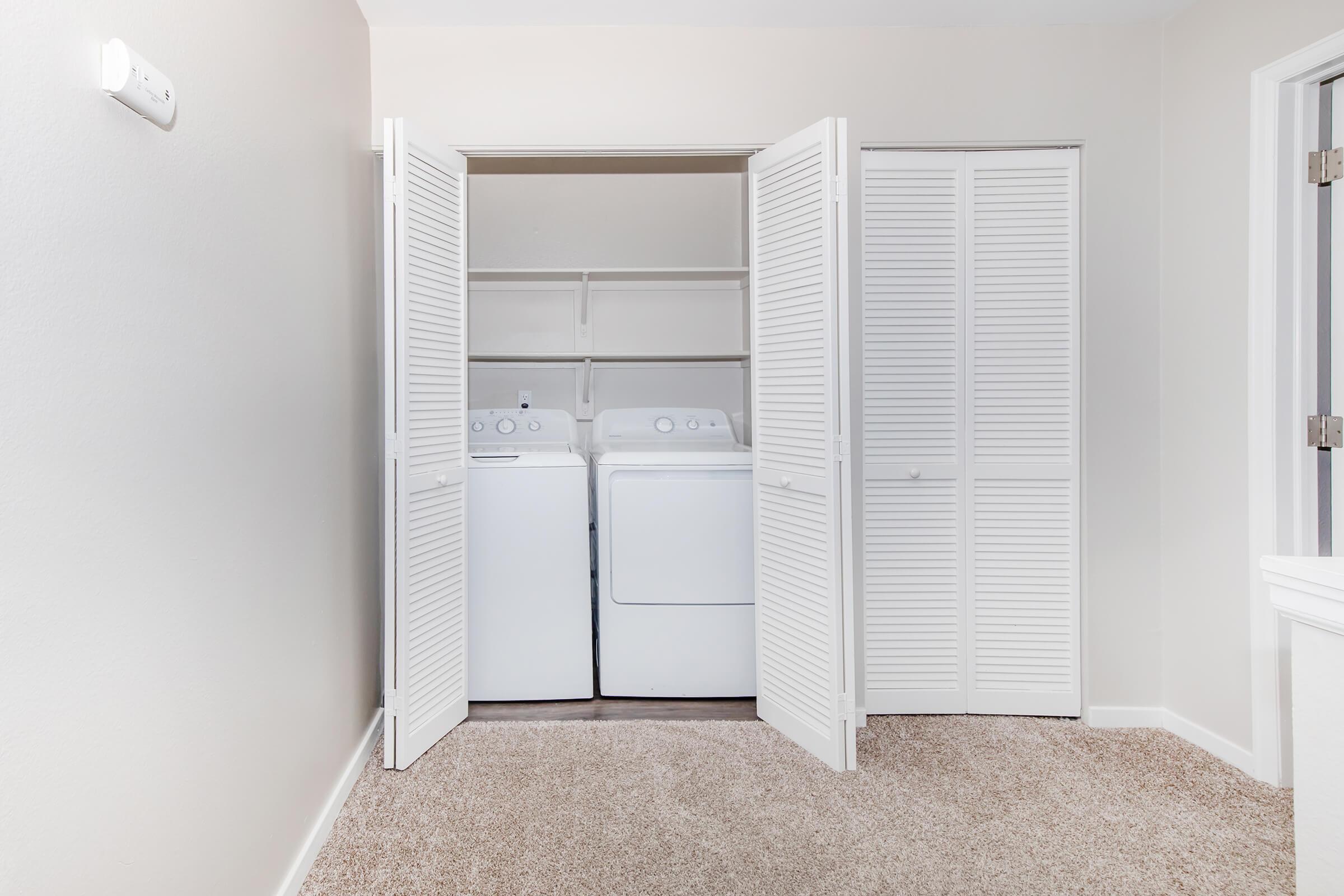 A laundry area with white front-loading washing machines and dryers situated in a closet. The doors are open, revealing shelves above the appliances. The space features light-colored walls and beige carpet, providing a clean and organized appearance. Natural light enters from a nearby window.