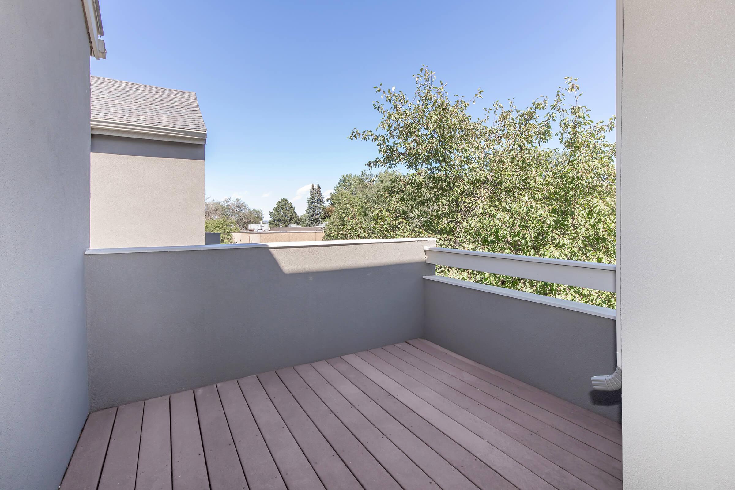 A small balcony with wooden flooring and a gray wall, overlooking trees and a clear blue sky. The surrounding area appears residential, with a glimpse of buildings and greenery in the distance.