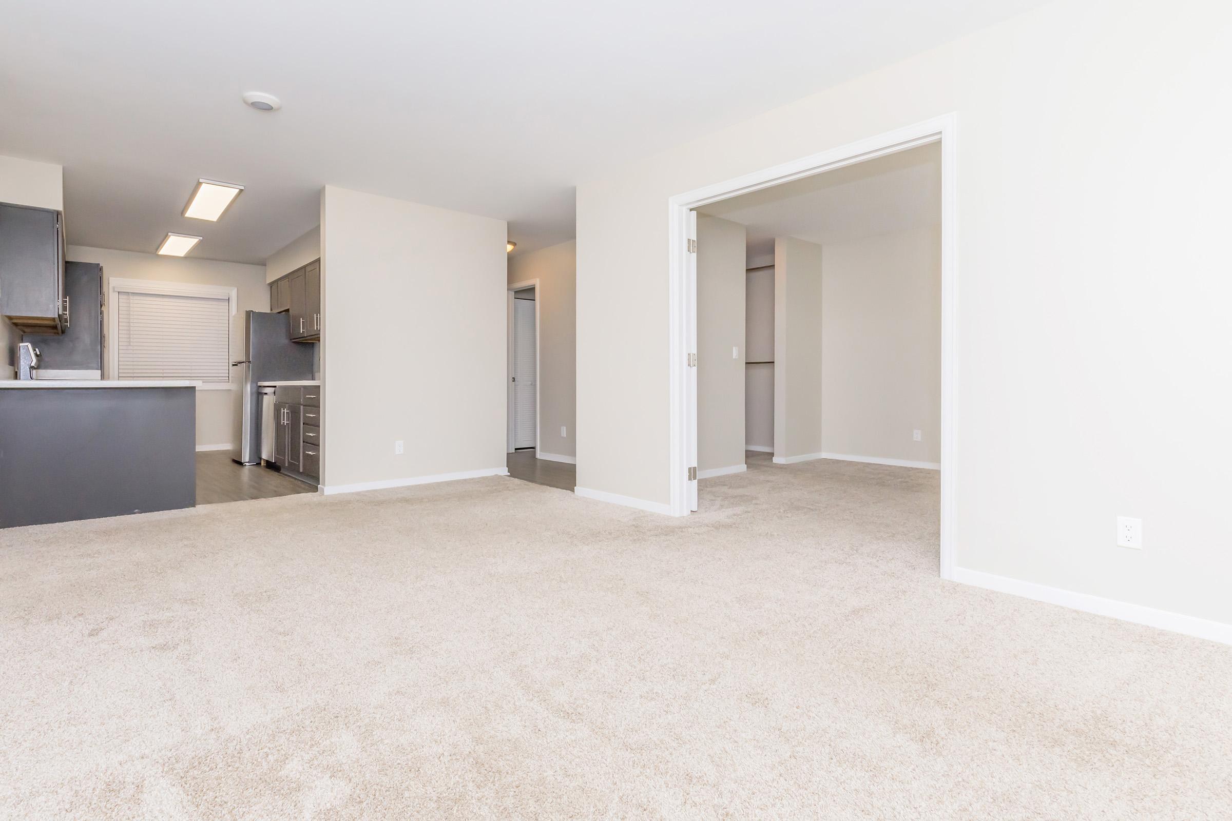 A spacious, empty living area with light beige carpet. The room features an open layout leading to a kitchen with modern appliances and light cabinetry. Walls are painted in neutral tones, contributing to a bright and airy atmosphere. Doors lead to adjacent rooms, enhancing the space's functionality.