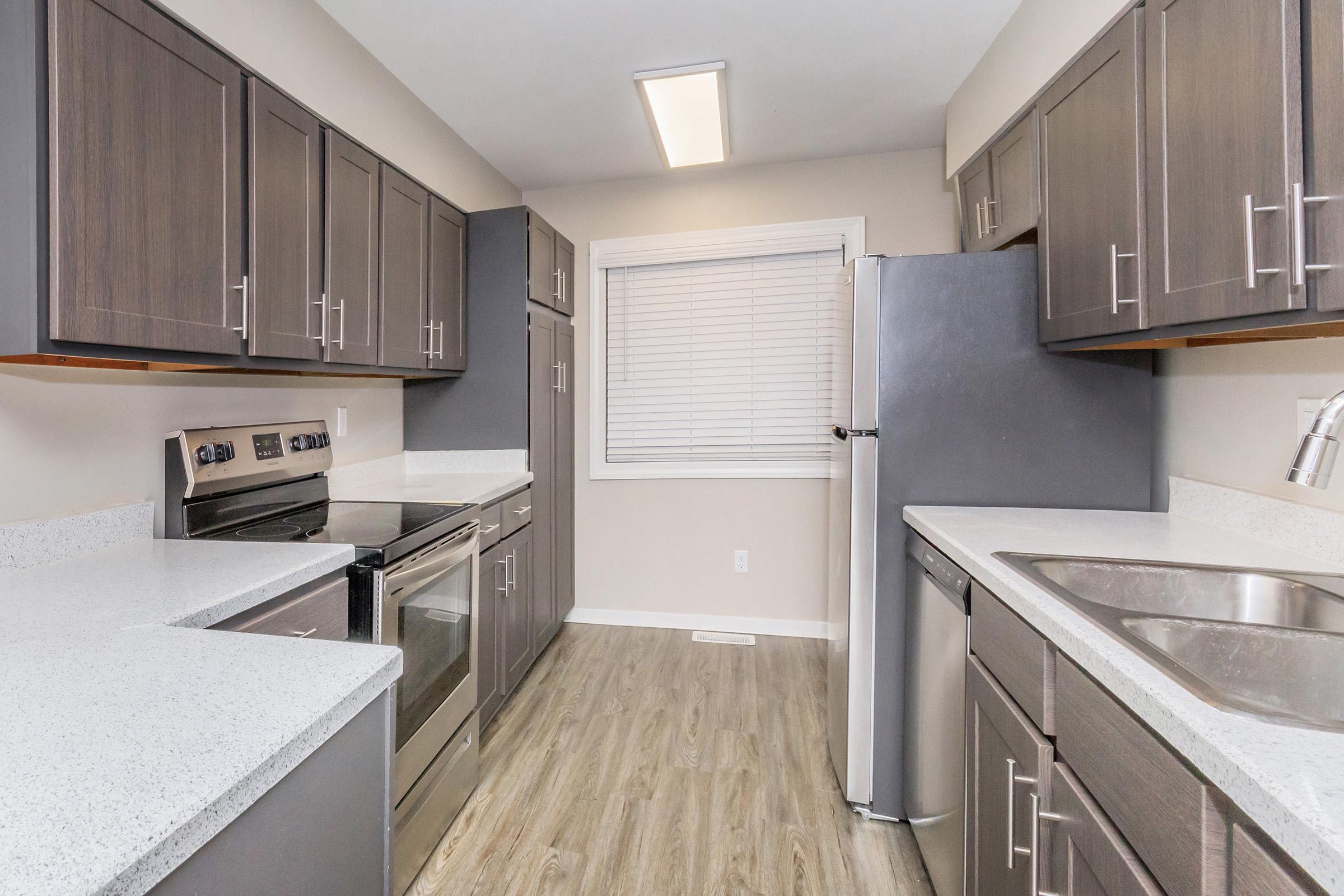A modern kitchen featuring dark cabinets, stainless steel appliances, and white countertops. The space includes an oven, a refrigerator, and a double sink, with a window providing natural light. The flooring is a light wood laminate, enhancing the contemporary design.