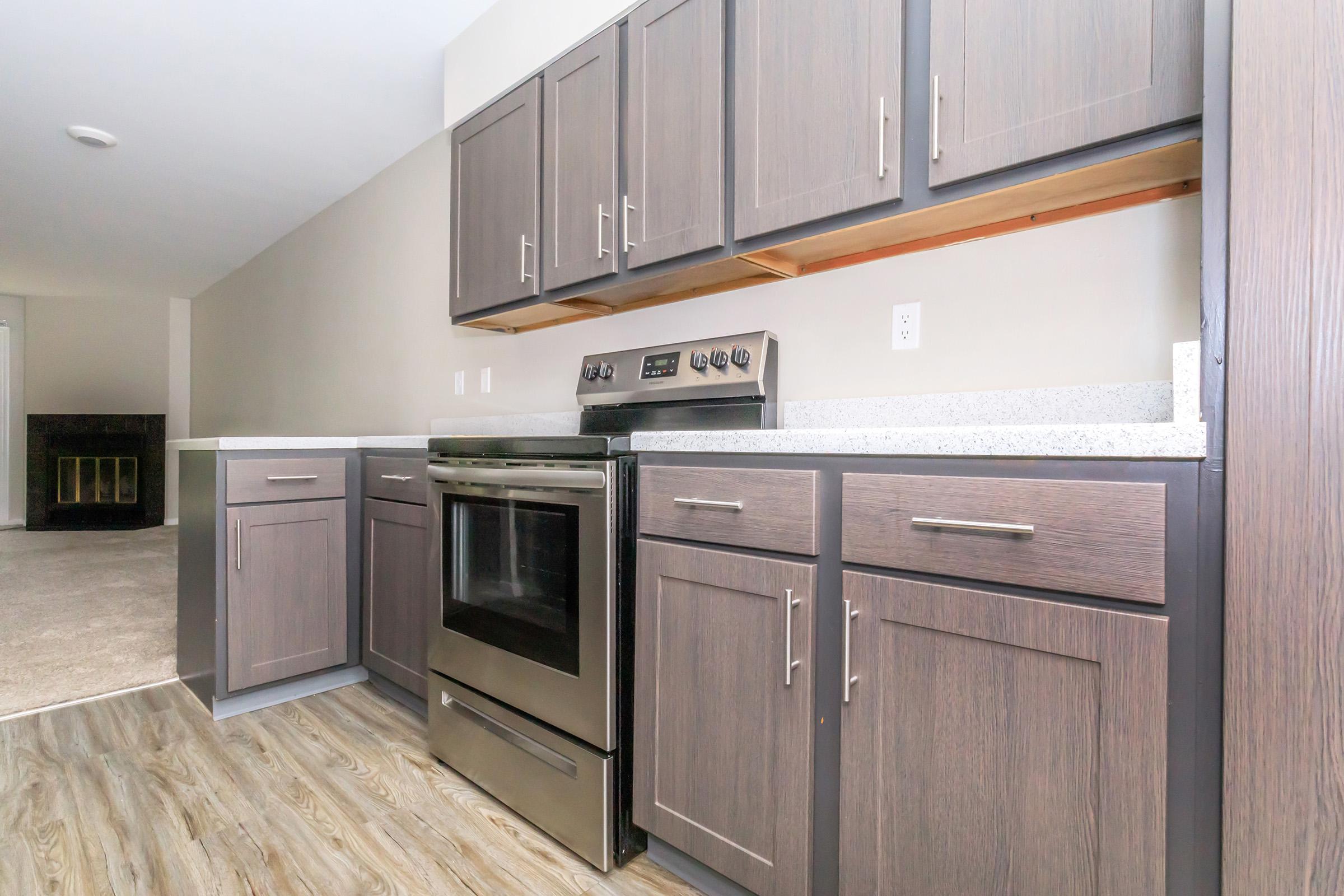 Modern kitchen with gray cabinetry, a black oven, and a granite countertop. The layout includes upper and lower cabinets, with a focus on a clean, contemporary design. The room has neutral-colored walls and a space that transitions to a carpeted area in the background.