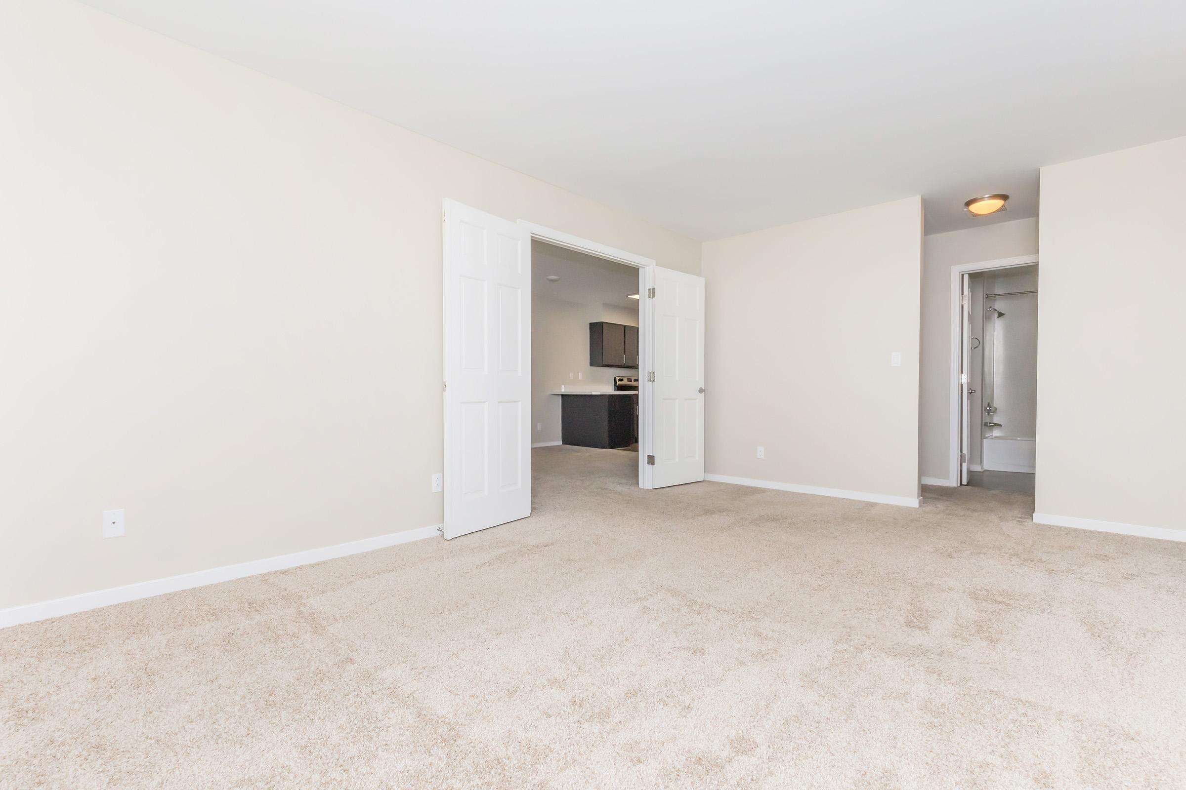 A spacious, empty room with beige walls and light-colored carpet. There are double doors leading to another area, and a doorway visible in the background that leads to a bathroom. Natural light illuminates the space, creating a bright and airy feel.