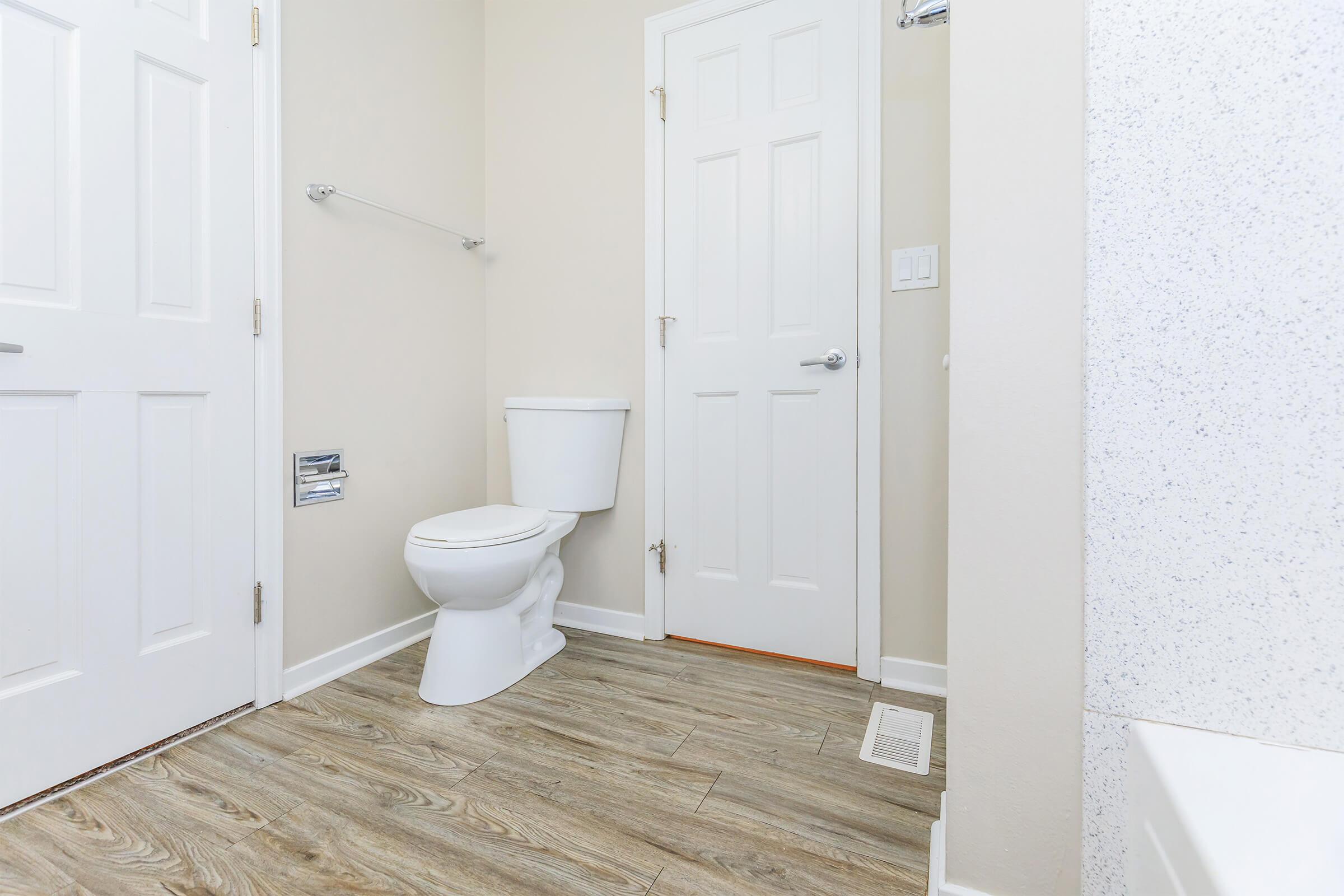 A clean bathroom interior featuring a white toilet, a closed door, and light-colored walls. The floor is covered with wood-like tiles, and there is a towel rack on the wall. A light fixture is visible, adding brightness to the space.