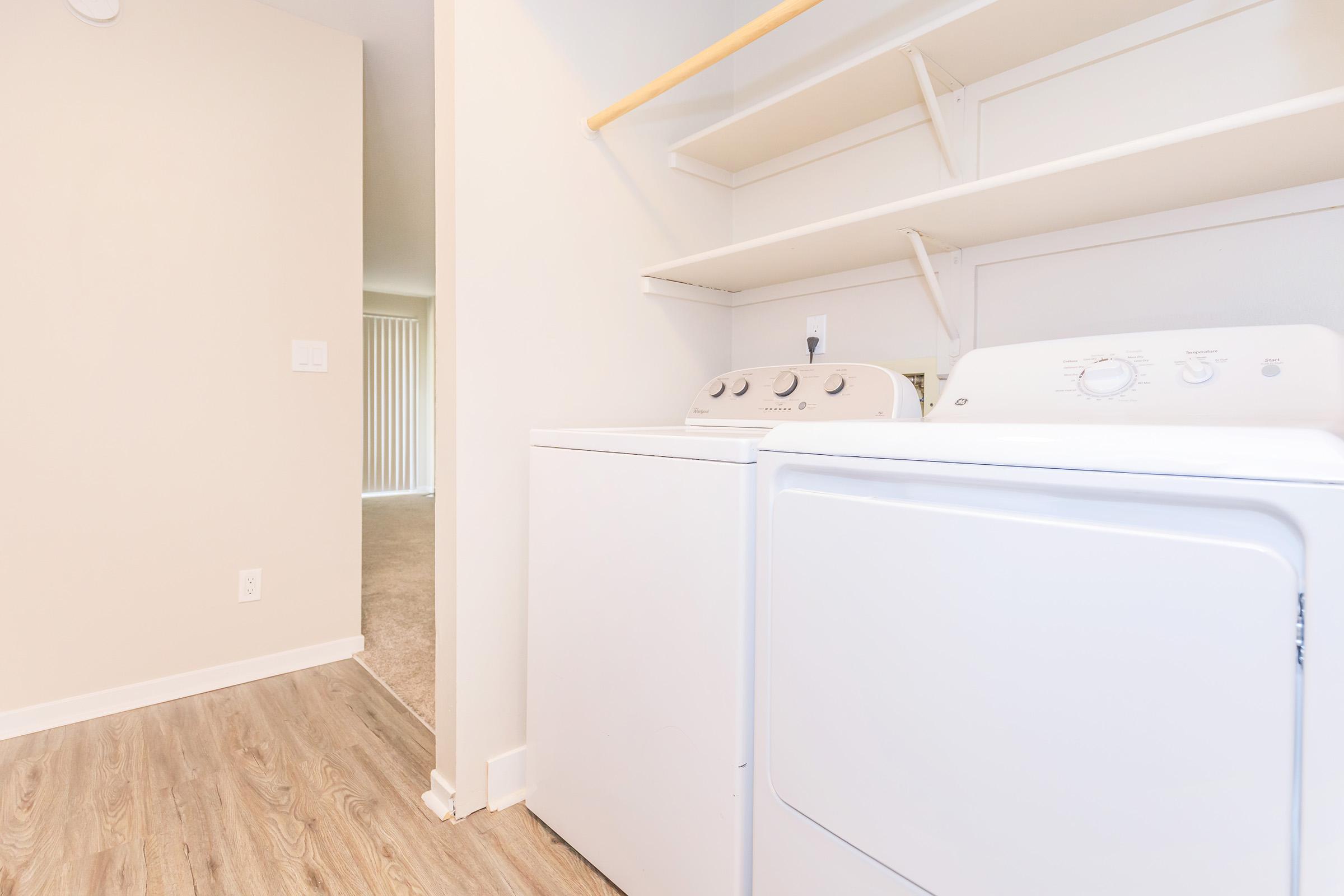 A laundry area featuring a white washing machine and dryer side by side, with shelves above for storage. The room has light-colored walls and a doorway leading to another area, emphasizing a clean and organized space. The flooring is wooden, adding warmth to the environment.
