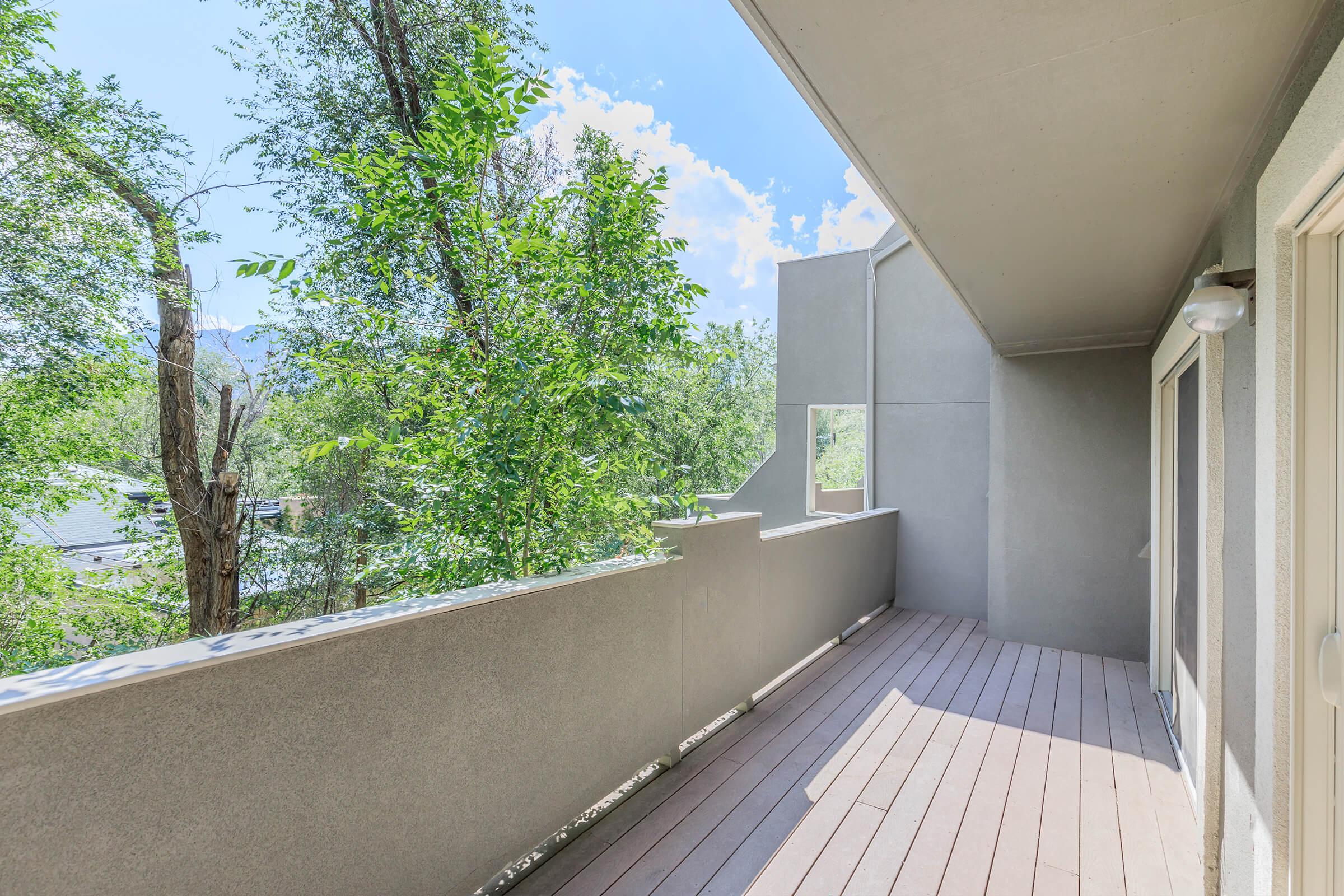 A spacious balcony with wooden flooring and gray walls, surrounded by lush green trees and a clear blue sky. The area features a railing and is partially shaded, creating a serene outdoor space ideal for relaxing. Sunlight filters through the trees, enhancing the peaceful atmosphere.