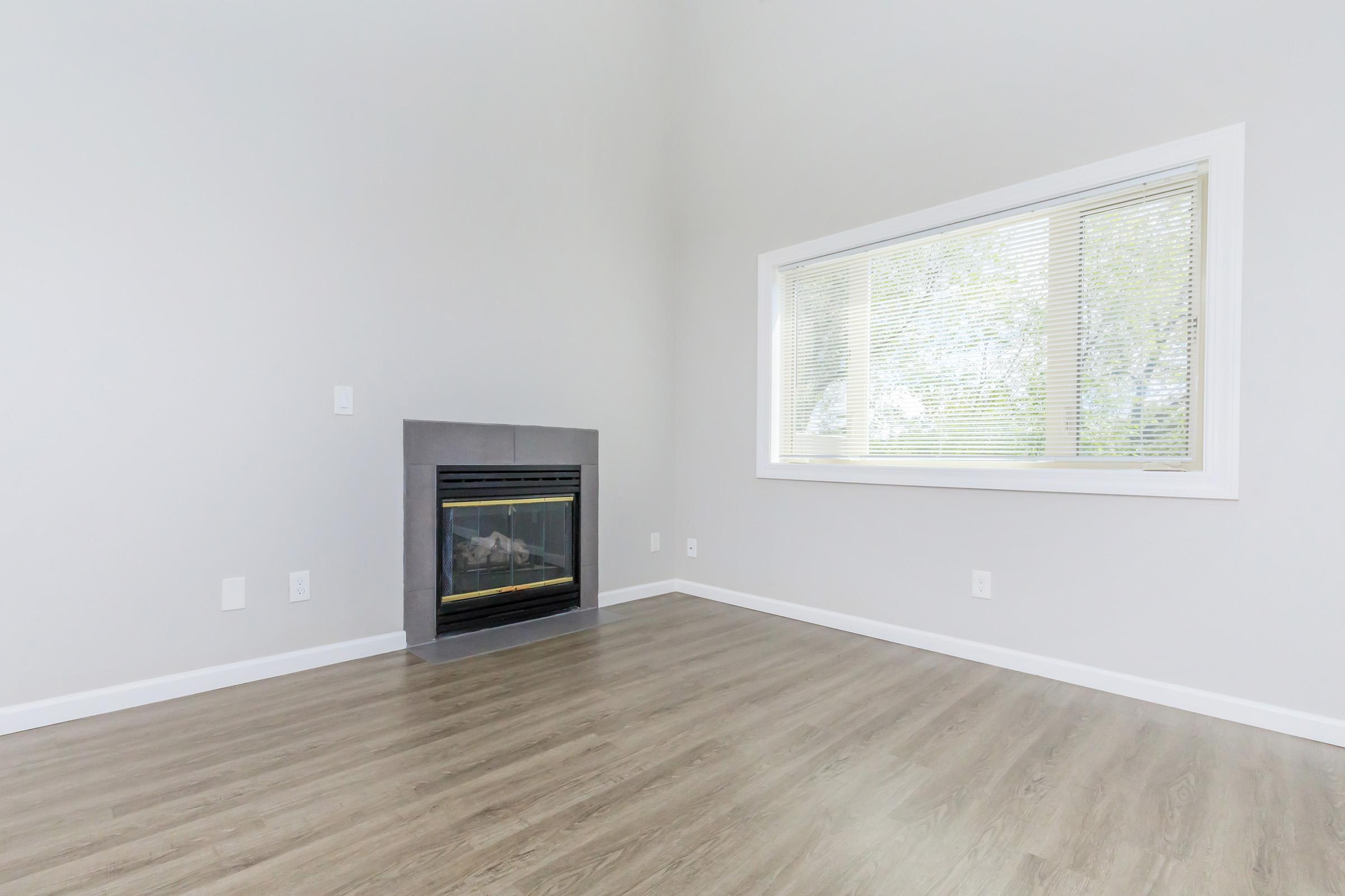 A spacious, empty living room featuring a modern fireplace on one wall, large window with blinds allowing natural light, and light-colored walls. The floor is made of laminate in a wood finish, creating a clean and minimalist aesthetic.