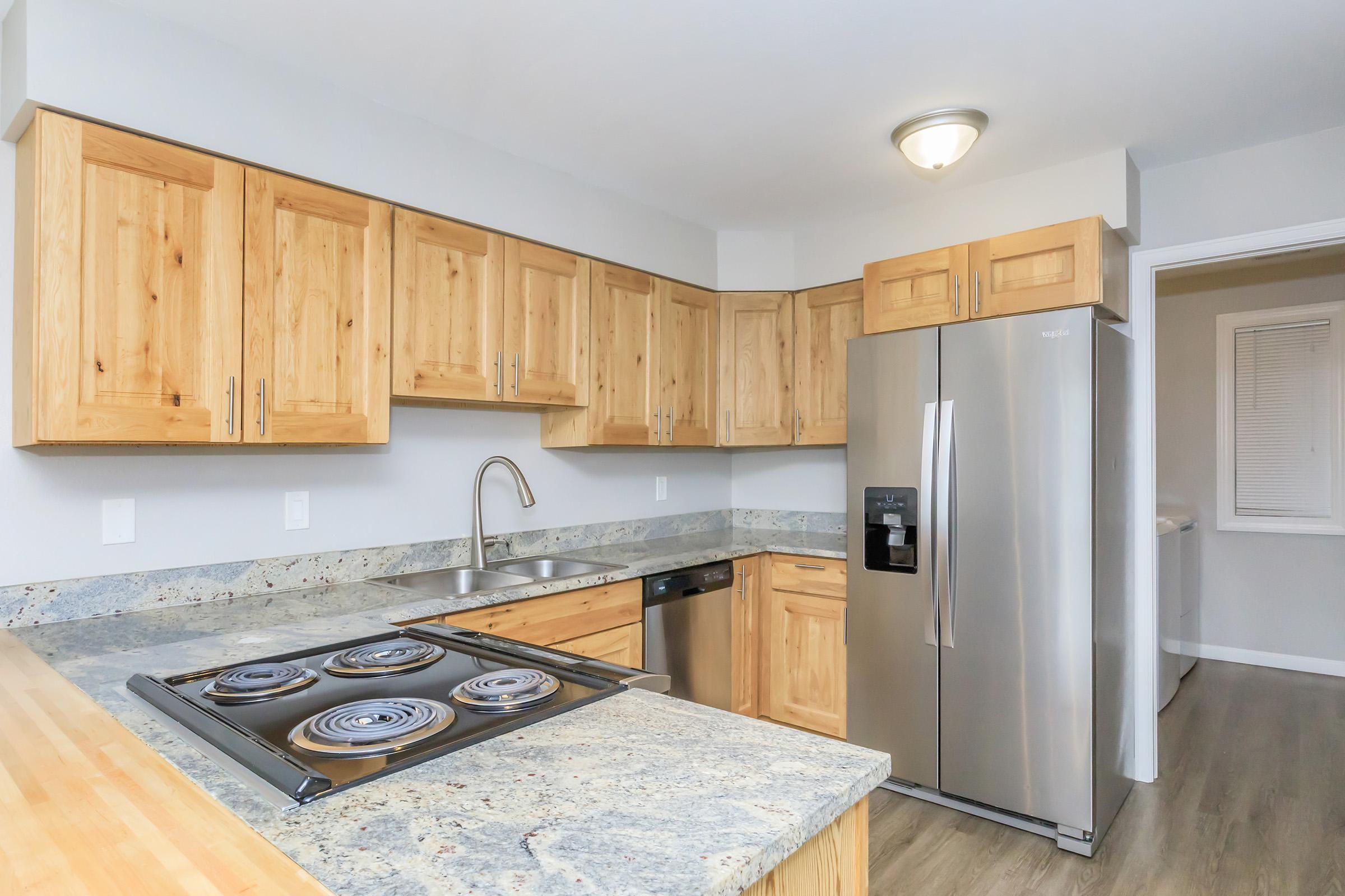 Modern kitchen featuring light wood cabinets, a stainless steel refrigerator, and a countertop with a built-in gas stove. The space is well-lit and includes a double sink, with a neutral color scheme of grey and natural wood tones.