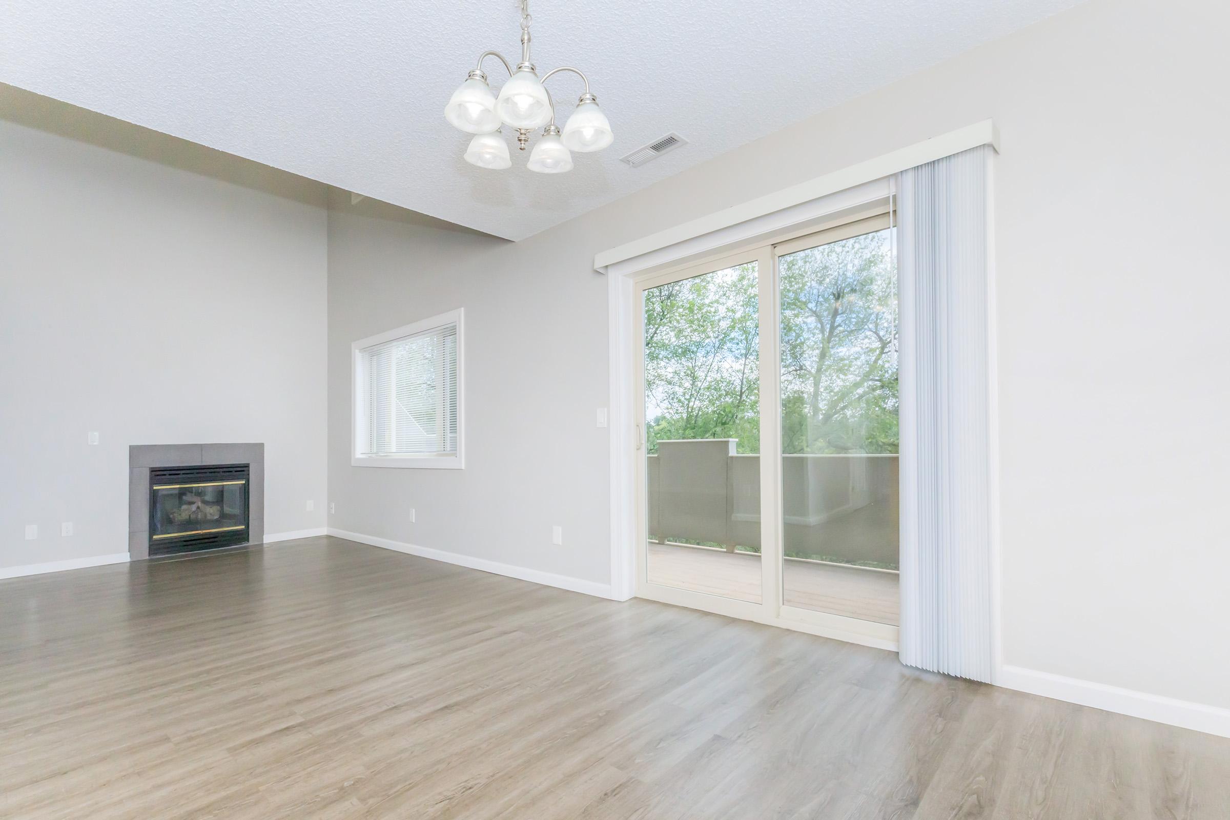 Spacious living room featuring large windows that provide natural light, a sliding glass door leading to a deck, and a modern fireplace. The walls are painted in a light color, and the floor is made of warm-toned wood. A simple chandelier hangs from the ceiling, adding elegance to the space.
