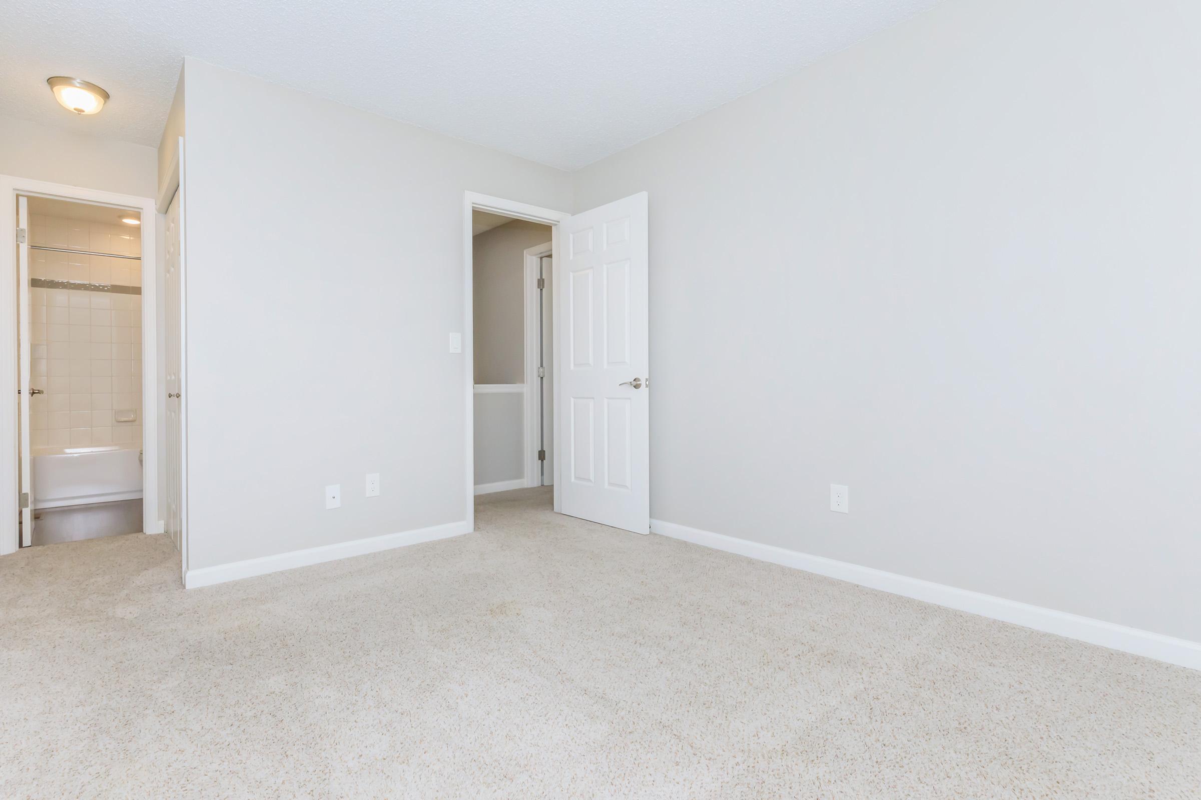 Empty room with gray walls and light beige carpet. The space includes a door leading to a bathroom in the background, and a second door on the left. The lighting is bright and the overall ambiance is clean and minimalistic. No furniture or decorations are present.