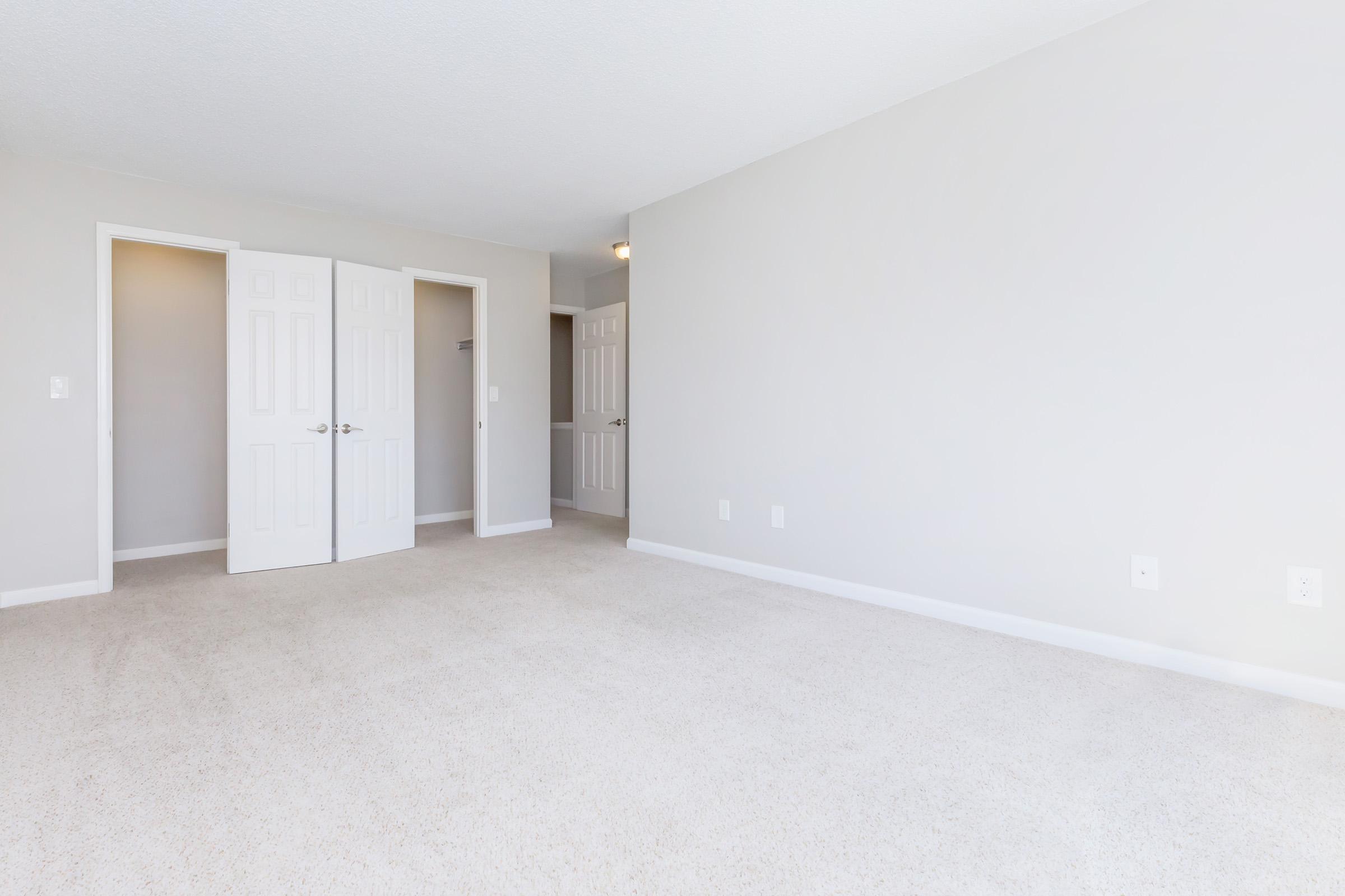 A spacious and bright room with light-colored walls and a plush carpet. Two white doors lead to adjacent spaces, and there are no furnishings, making it look empty and open. Natural light fills the area, creating a calm and inviting atmosphere.