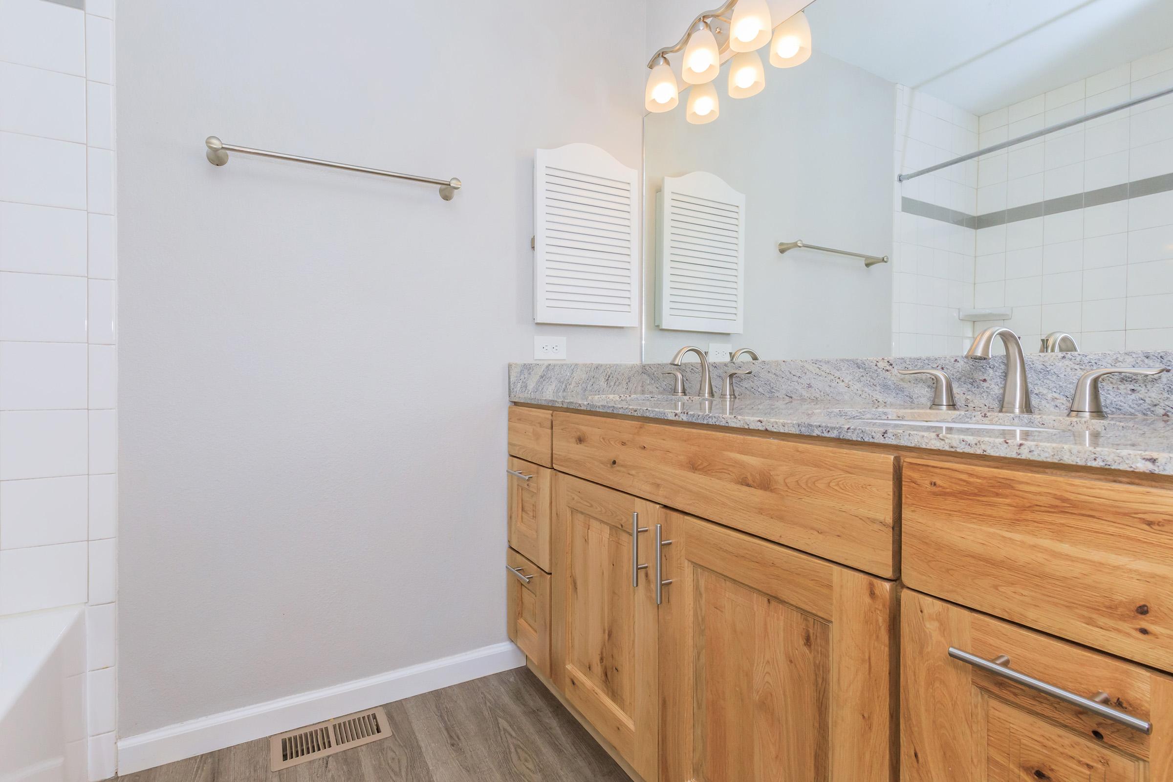 A modern bathroom with a light gray wall and wooden cabinets. The countertop is made of gray stone, featuring dual sinks with sleek faucets. There are two round light fixtures above the mirrors, and a towel bar is mounted on the wall. The flooring is a light wood-like material, and a bathtub is visible in the corner.