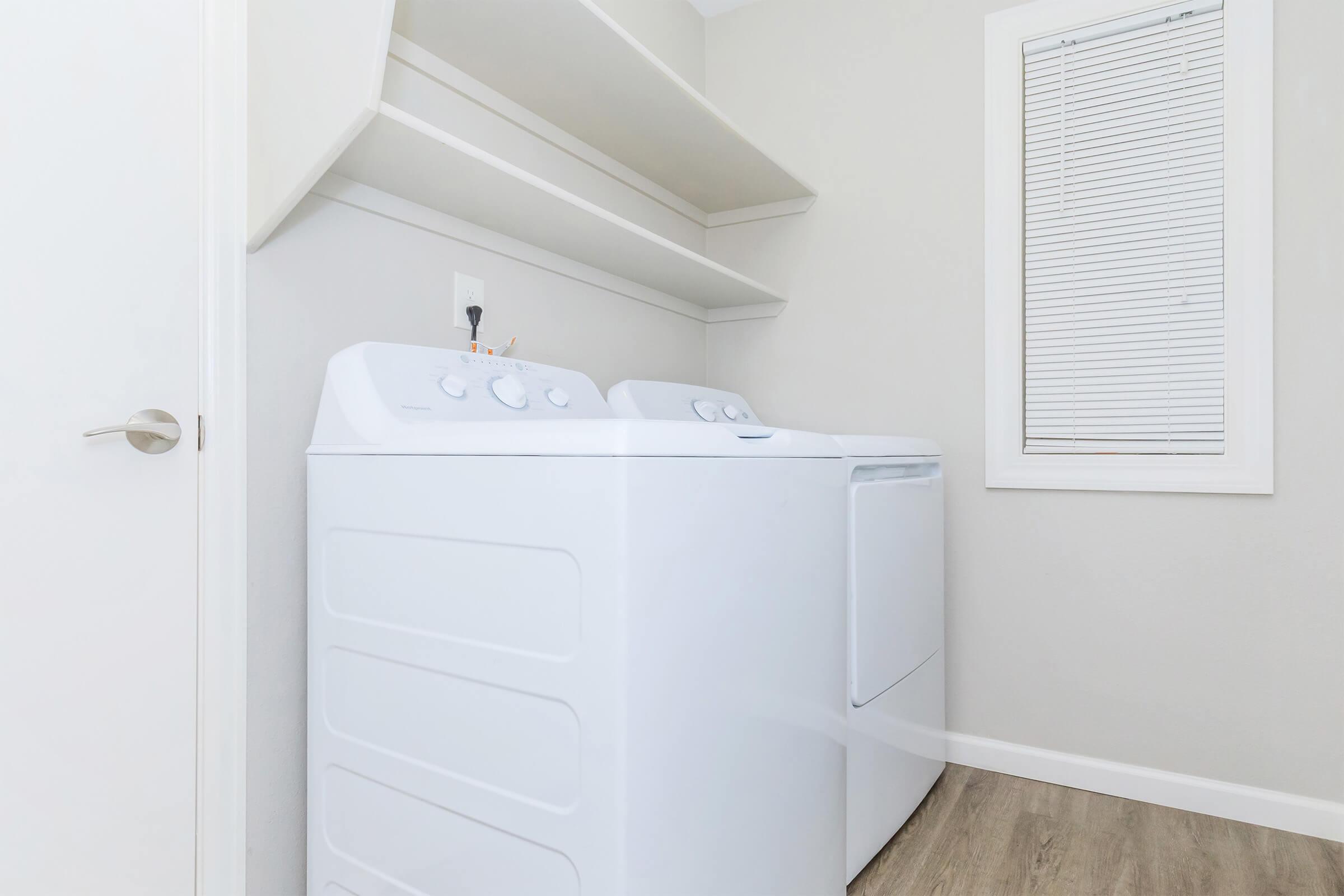 A clean laundry room featuring a white washer and dryer side by side, with two open shelves above them. A window with closed blinds is visible on the right side, providing natural light to the space. The floor is a light wood finish, contributing to a bright and modern aesthetic.