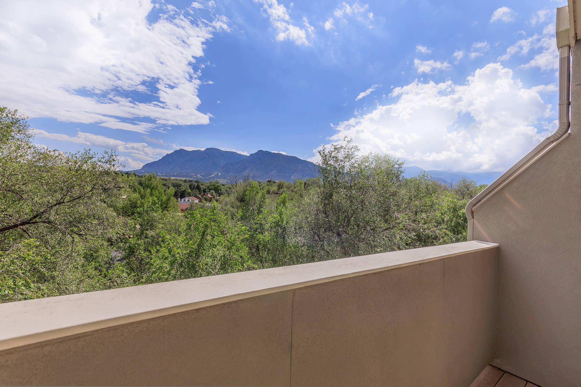 A scenic view from a balcony overlooking lush green trees and a mountainous landscape under a partly cloudy sky. The mountains are prominent in the background, contrasting with the vibrant greenery in the foreground.