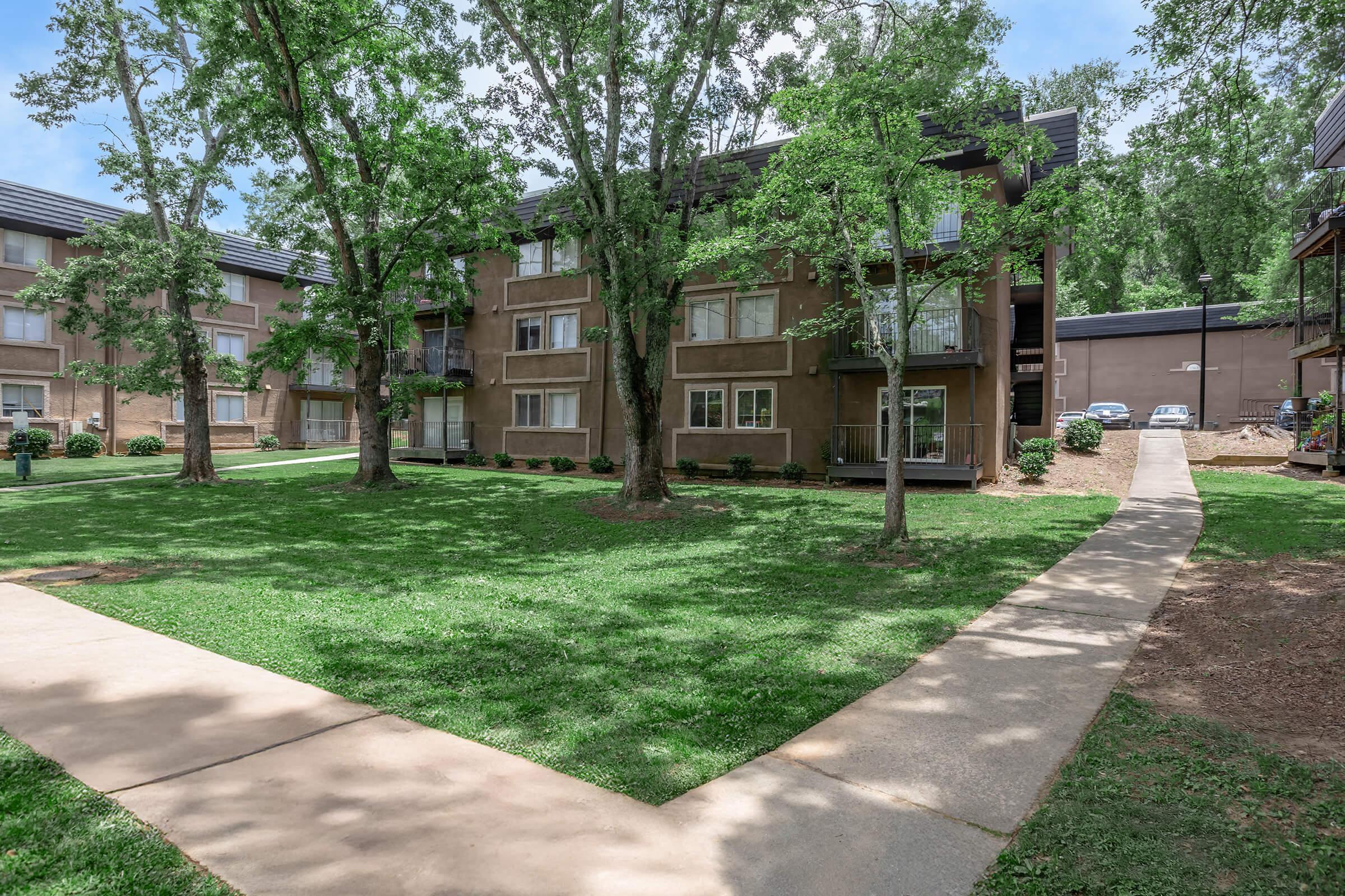 A view of a multi-story apartment building surrounded by green grass and trees. A paved walkway leads through the lawn, with additional parking visible in the background. The sky is partly cloudy, creating a pleasant residential environment.