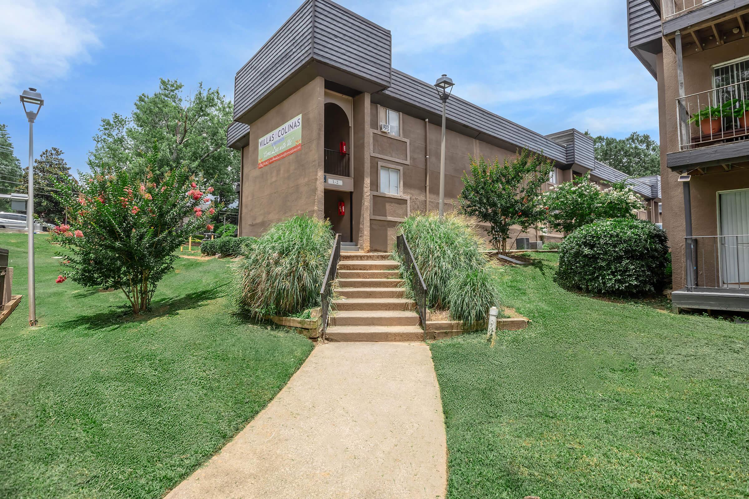 A view of an apartment building with a walkway leading to the entrance. Surrounding greenery includes shrubs and small trees, with a lamp post on the left. The building features a sign with the name of the residence, and there are balconies on the upper floors. The sky is clear and blue.