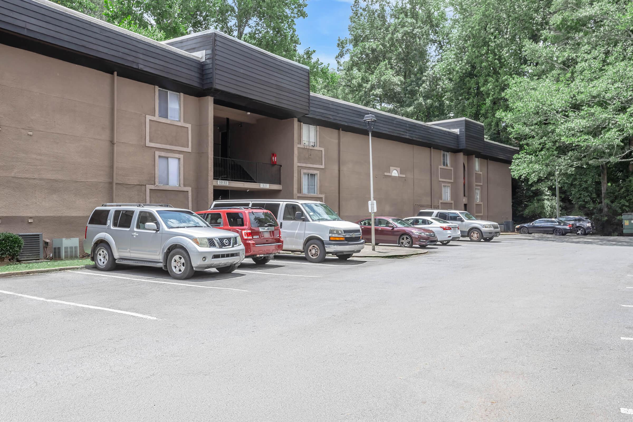 A parking lot in front of a two-story residential building surrounded by trees. Several vehicles, including SUVs and a van, are parked in designated spaces. The sky is clear with a few clouds, and the building features a modern design with balconies.