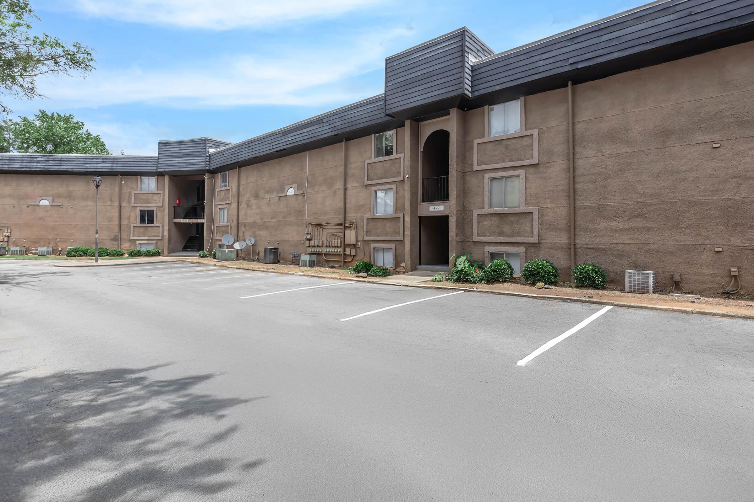 A view of a two-story apartment building with a beige exterior. It features multiple windows and balconies, surrounded by neatly trimmed shrubs and a paved parking lot with empty parking spaces. The sky is clear, adding to the bright atmosphere of the scene.