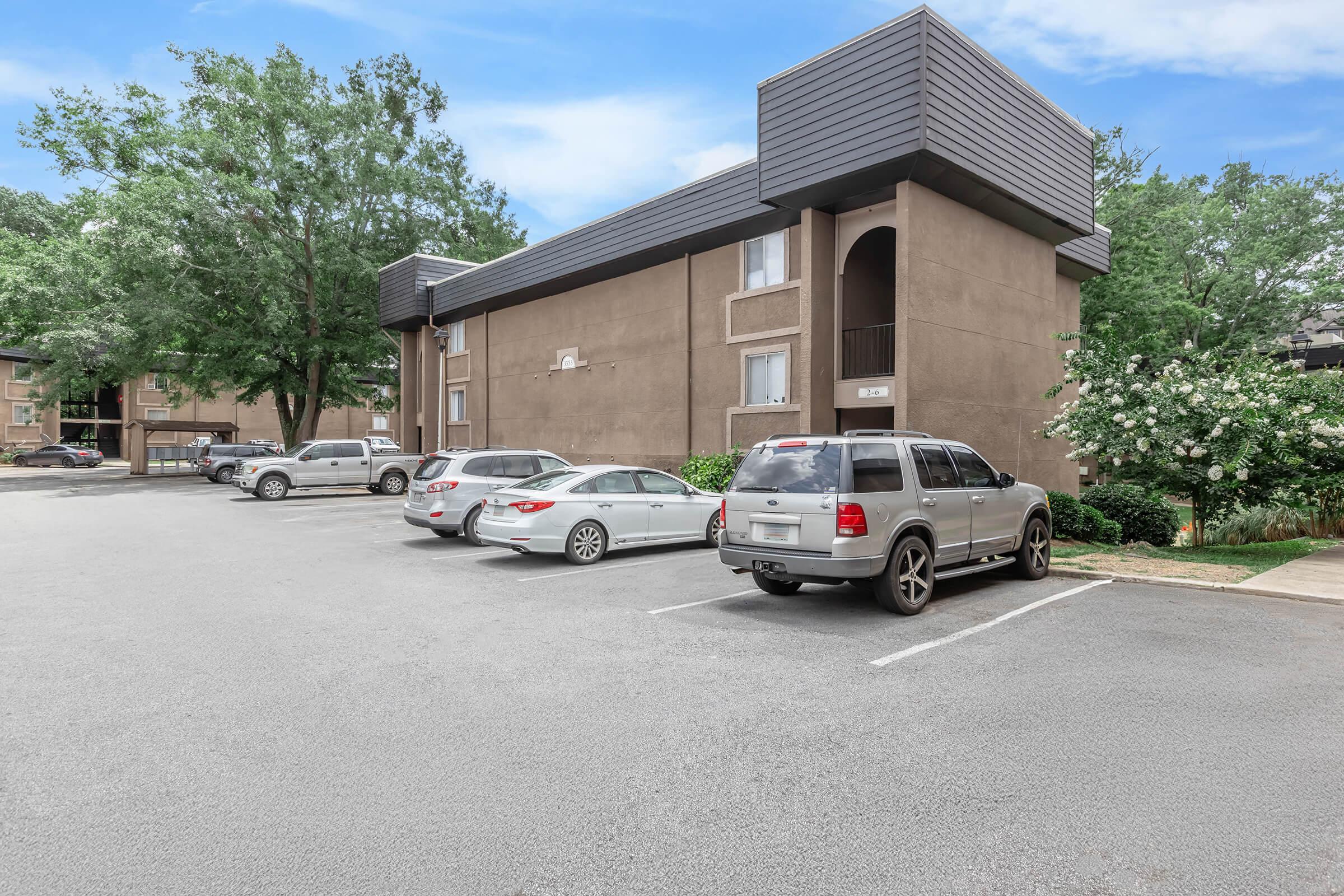 A view of an apartment complex with a parking lot in the foreground. Several cars are parked, and the building has a modern design with a dark exterior and balconies. Lush greenery is visible nearby, under a clear blue sky.