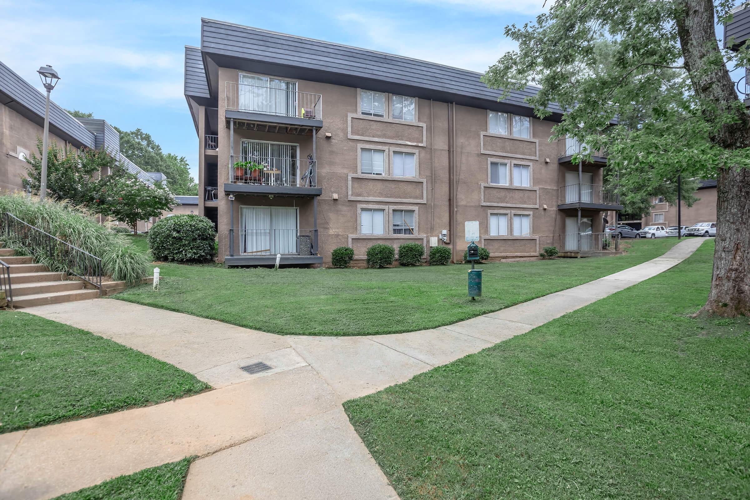 A view of a multi-story apartment building surrounded by green lawns and trees. The building features balconies with some plants, a sidewalk leading up to the entrance, and a lamp post nearby. The setting appears calm and well-maintained, suitable for residential living.