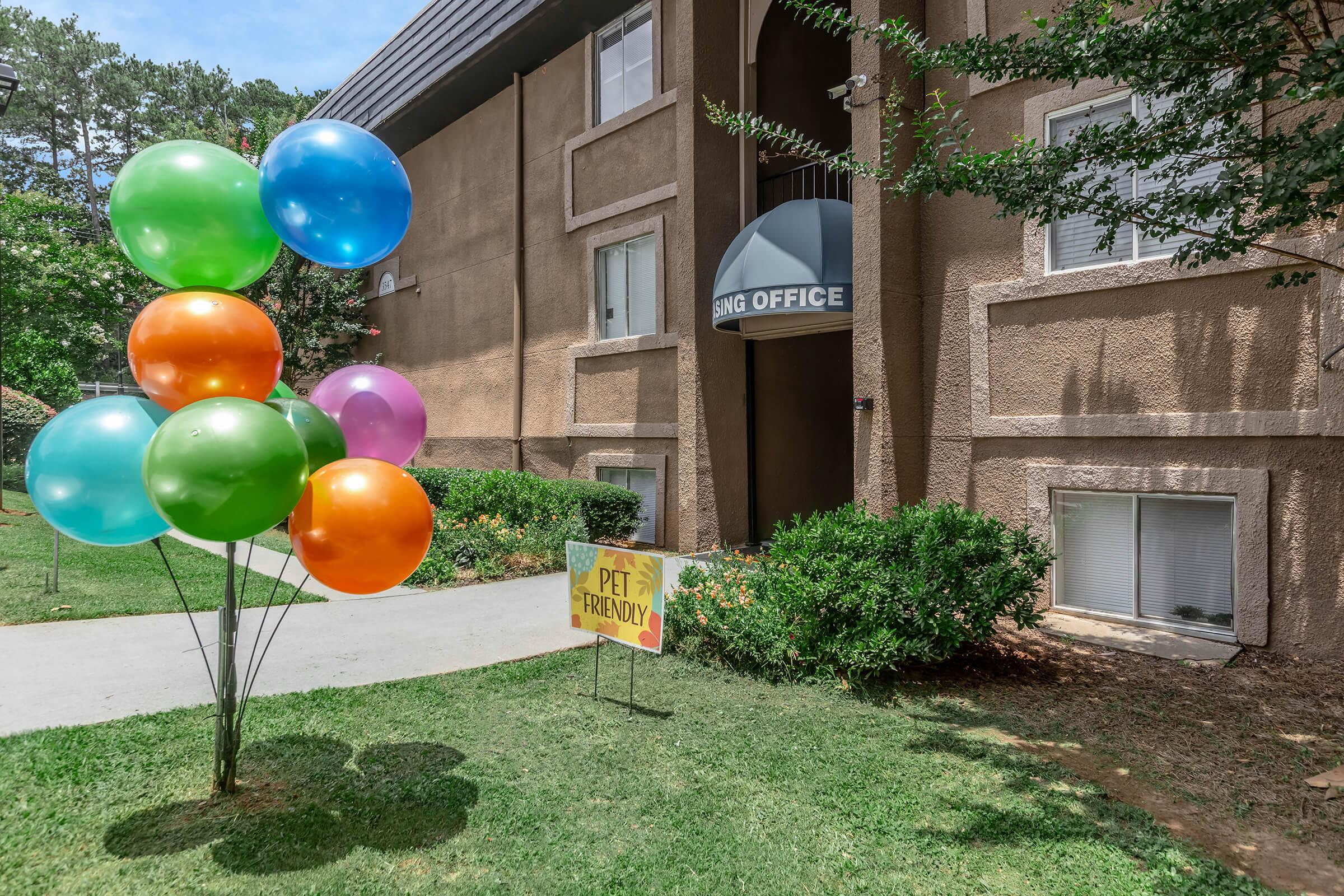 A welcoming apartment leasing office surrounded by greenery. Colorful balloons in various shades of blue, green, orange, pink, and purple are tied together and stand in front of the building. A sign reading "PET FRIENDLY" is prominently displayed on the lawn. Bright and inviting atmosphere.