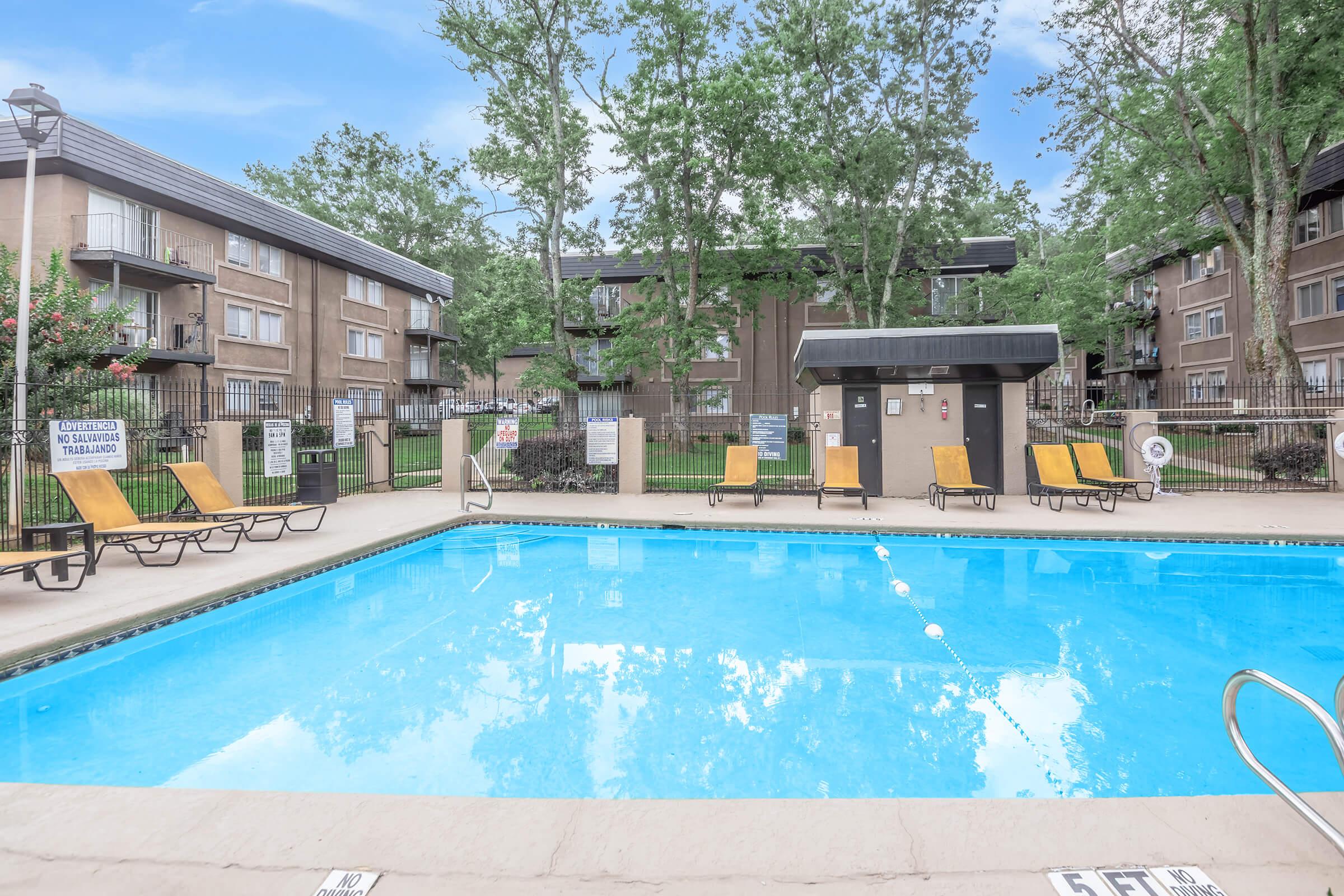 A clear swimming pool with lounge chairs on the side, surrounded by lush greenery and residential buildings in the background. The scene is bright and inviting, perfect for relaxation. There are signs around the pool area, and the sky is blue.