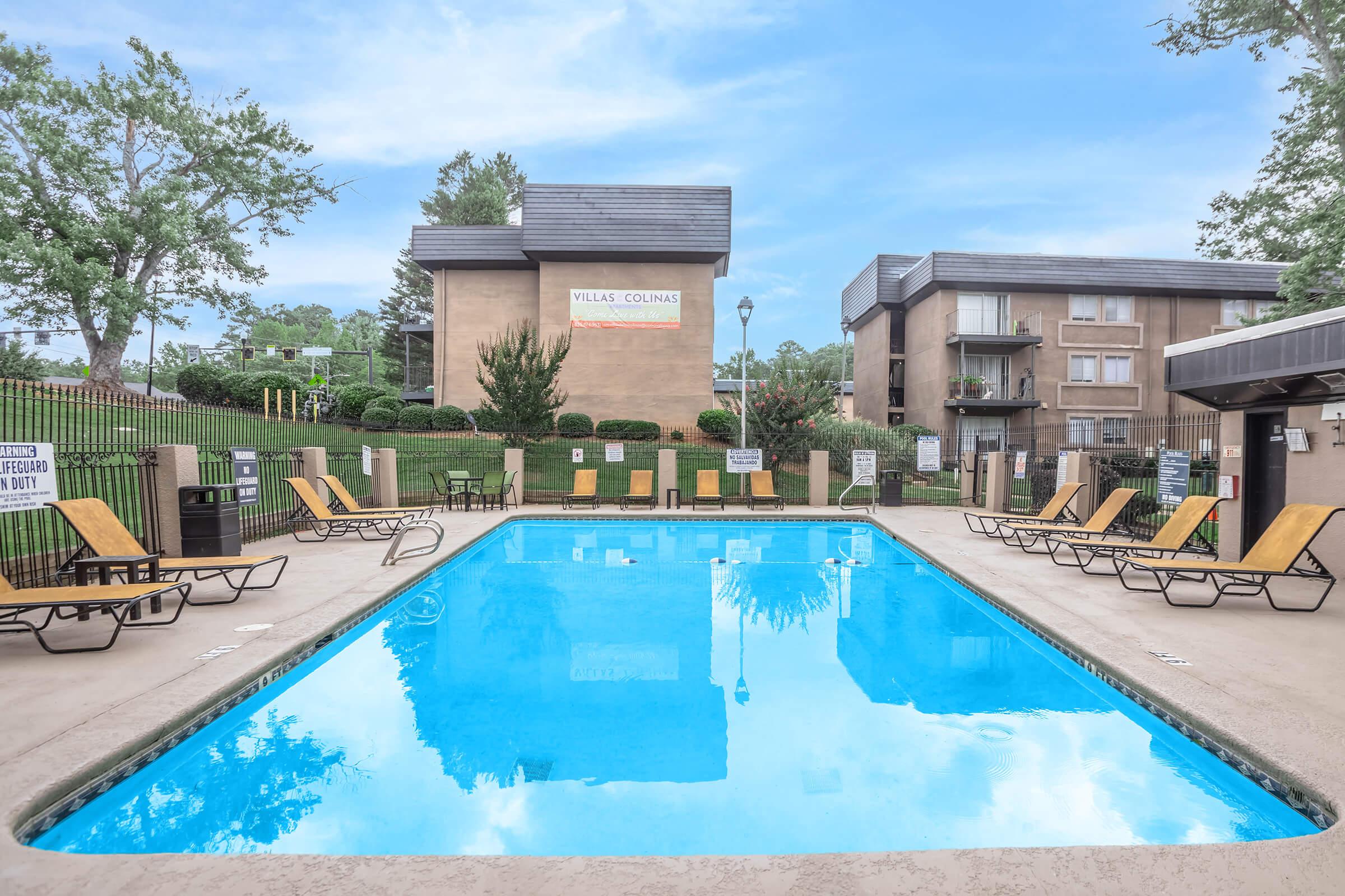 A clear blue swimming pool surrounded by lounge chairs in a residential complex. Trees and a building are visible in the background, with a sign for "Villas Colonies" prominently displayed. The scene is bright and inviting, showcasing a relaxing outdoor space.