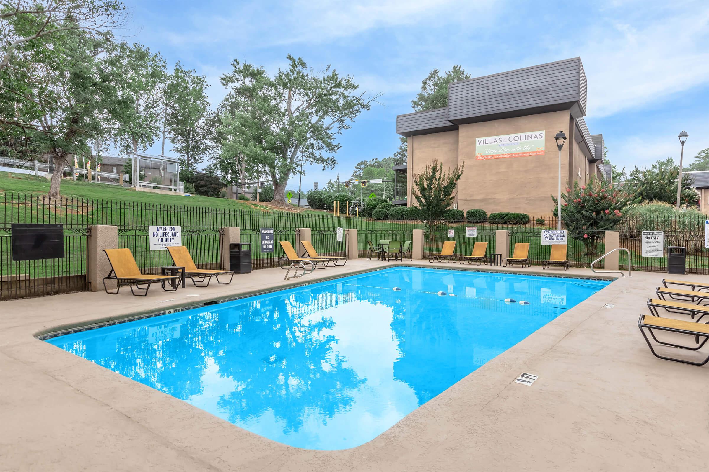A clear blue swimming pool surrounded by lounge chairs. Trees and a building are visible in the background, with signs indicating pool rules. The scene is sunny and inviting, ideal for relaxation and leisure.