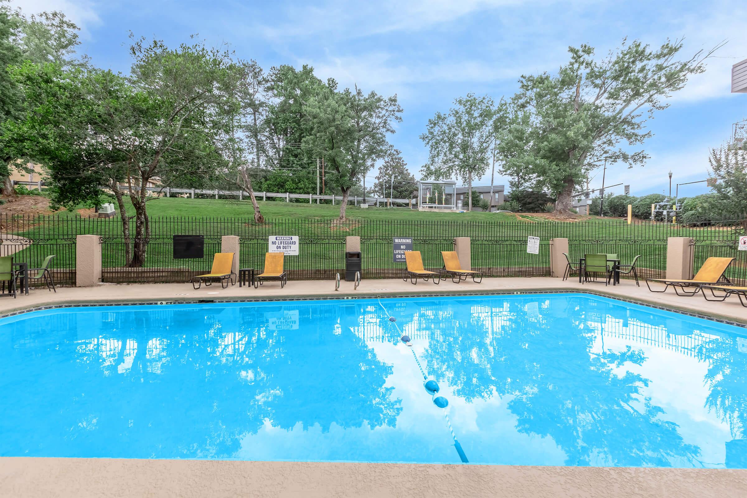 A clear blue swimming pool surrounded by lounge chairs, with green trees in the background and a grassy area. A fence encloses the pool area, and there are several warning signs visible. The sky is partly cloudy, creating a serene outdoor setting.