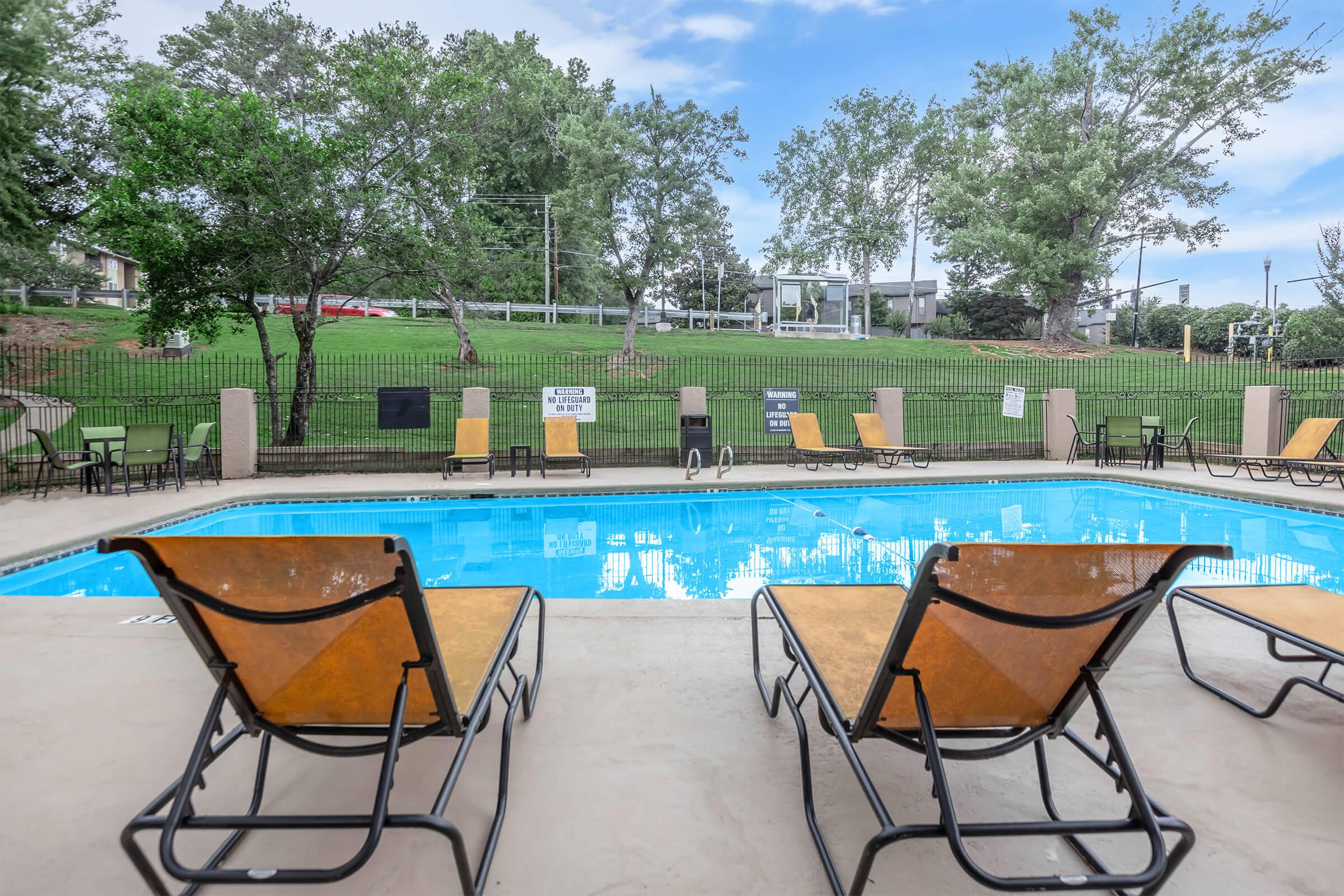 A clear swimming pool surrounded by lounge chairs and green grass. Trees are visible in the background, along with a fence. The scene appears inviting and relaxing, perfect for enjoying outdoor activities.