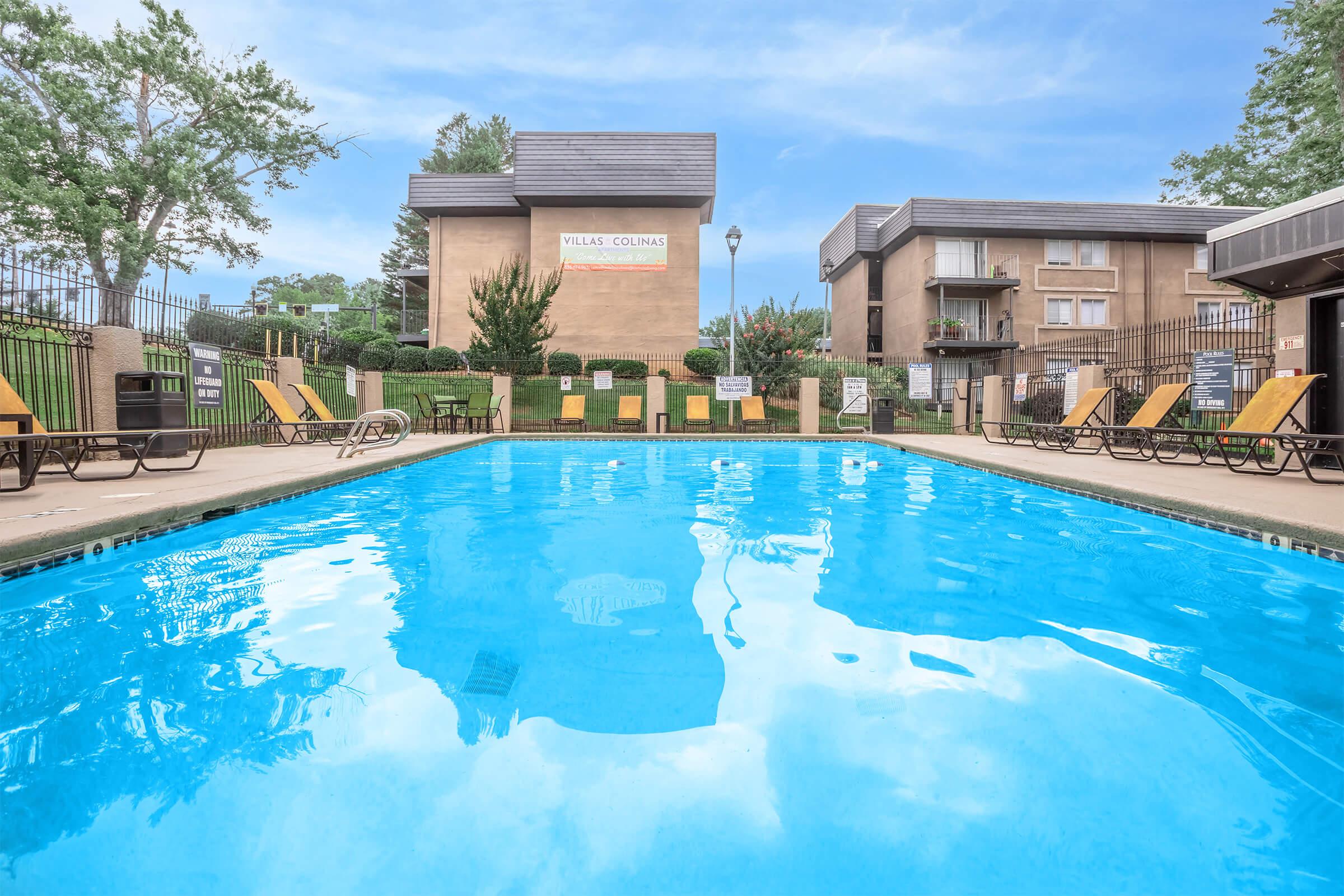 A clear swimming pool in the foreground, surrounded by lounge chairs. The background features a well-maintained apartment complex with greenery and trees. There are signs around the pool area and a lamp post. The sky is blue with a few clouds, creating a serene and inviting atmosphere.